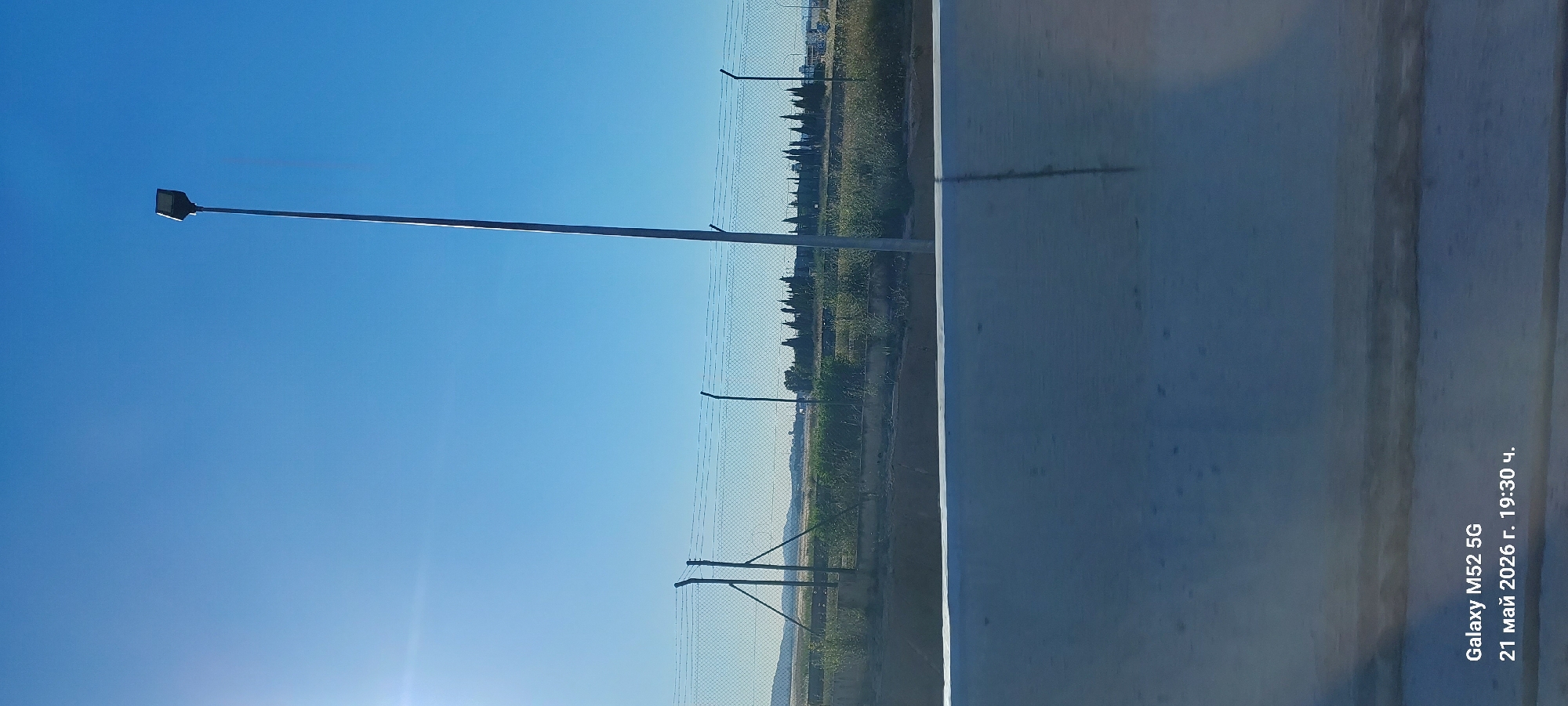 The image depicts an outdoor scene with a clear blue sky. A tall street light with a modern-looking fixture is visible on the left side of the frame. In the background, a chain-link fence runs along the horizon, separating the foreground from a grassy area with scattered vegetation and trees. The right side of the image shows a white surface with some streaks or stains. The location is possibly in or near Quart de Poblet, Spain. There are no people visible in the image. The photo was taken on a Galaxy M52 5G on 21 May 2026.