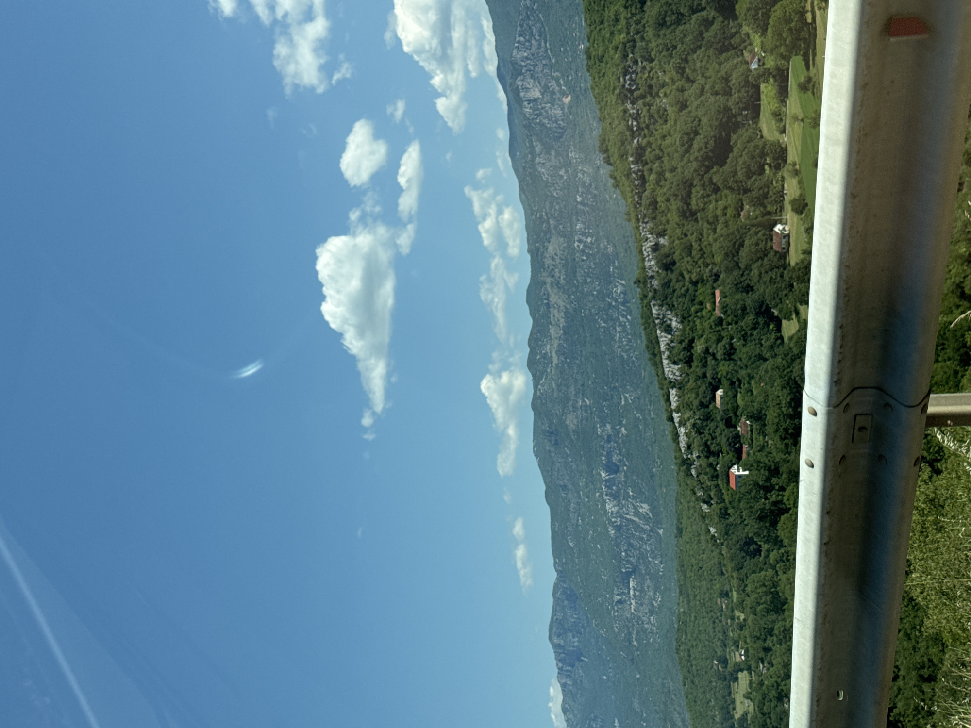The image shows a landscape with mountains in the background and lush green trees in the foreground. There are a few small houses scattered among the trees, suggesting a rural setting. A metal guardrail runs along the bottom of the image, indicating that this view might be from a road or an elevated viewpoint. The sky is blue with a few scattered white clouds. The scene appears to be captured during the daytime under clear weather conditions.