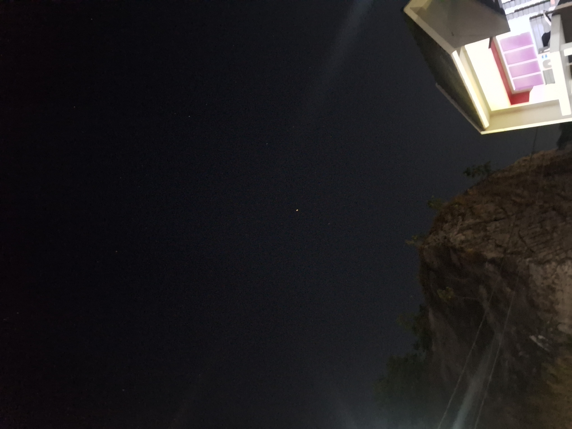 The image shows a dark night sky with a few stars visible. In the lower right quadrant of the image, a building with a white exterior and red-tinted windows is illuminated. To the left of the building, a rocky embankment with some vegetation is visible. Power lines are strung across the embankment. The scene appears to be an outdoor area in Samarinda, Indonesia, possibly near a construction site or a residential area at night. There are no people or animals in the image.