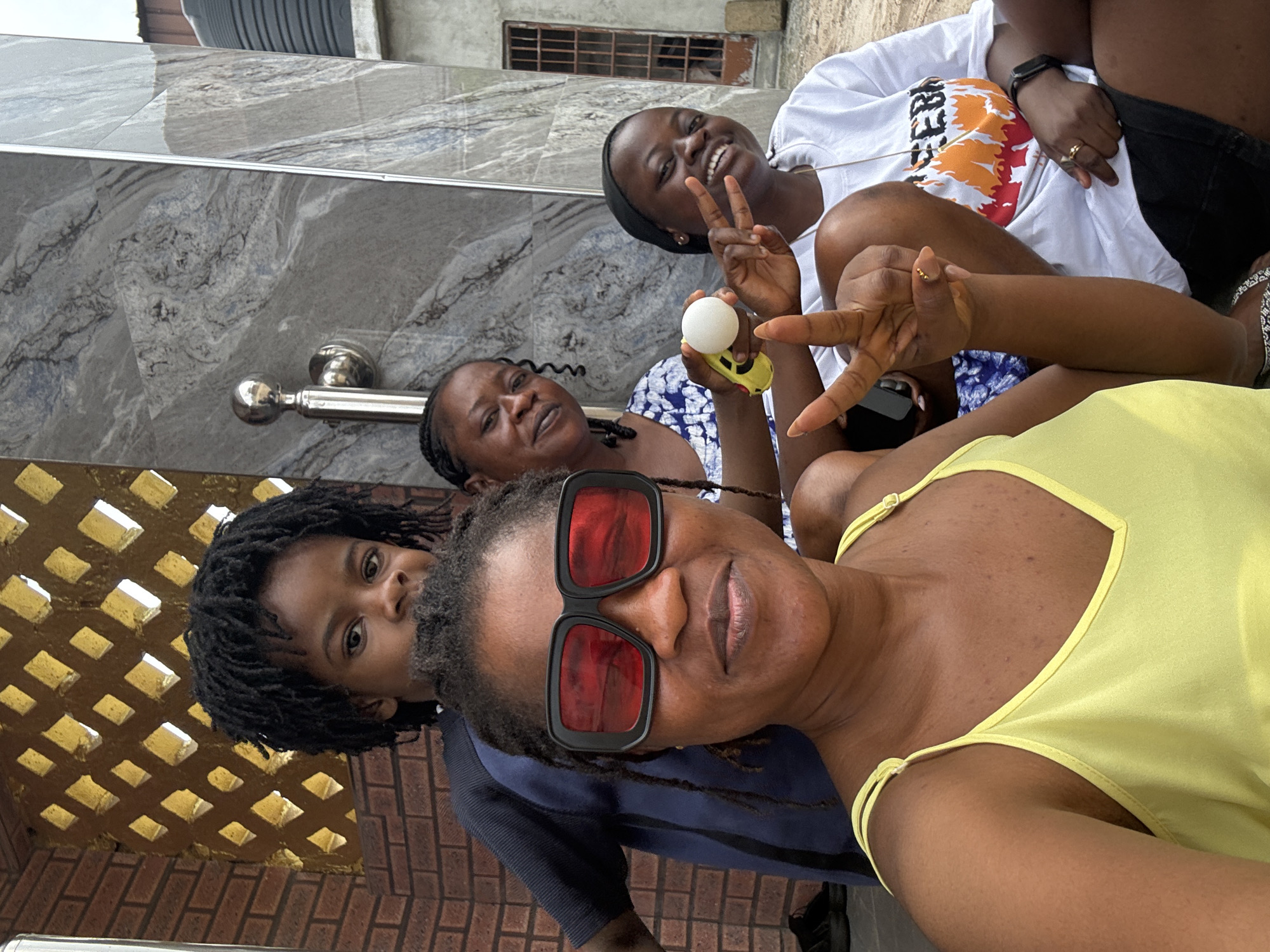 In the city of Ajara, Nigeria, a group of four individuals poses for a selfie in an outdoor setting. The foreground is dominated by a woman wearing a yellow tank top and large red-tinted sunglasses. Behind her, a child with dark dreadlocks looks directly at the camera. To the right of the child, another woman in a blue and white patterned top holds a small white object. The fourth person, on the far right, wears a white t-shirt and black shorts, and flashes a peace sign with their fingers. The background features a building with a decorative facade of brick and marble-like stone, a metal railing, and a barred window. The ground appears to be sandy or dusty. The overall scene suggests a casual gathering.