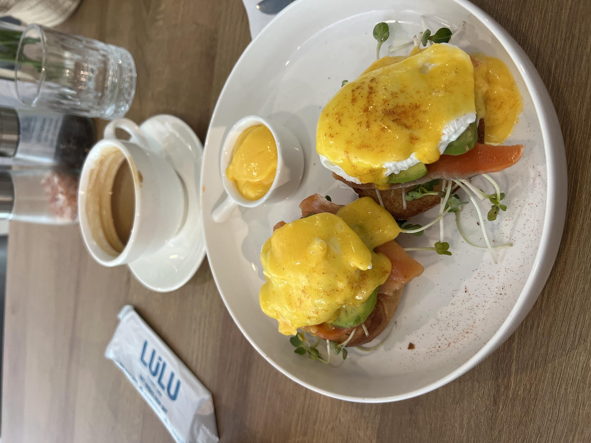 This is a medium-shot, eye-level photograph of a plate of food and a cup of coffee. The photograph appears to have been taken in Bac Nha Trang Ward, Vietnam. The main subject is a white plate containing two eggs benedict with salmon and avocado, garnished with sprouts, accompanied by a small ramekin of hollandaise sauce. The eggs benedict sit on top of toasted bread. To the left, there is a white coffee cup with coffee and a saucer, a glass, salt and pepper shakers, and a wrapped item with "LULU" printed on it. The surface appears to be a wooden table. The lighting is bright and natural.