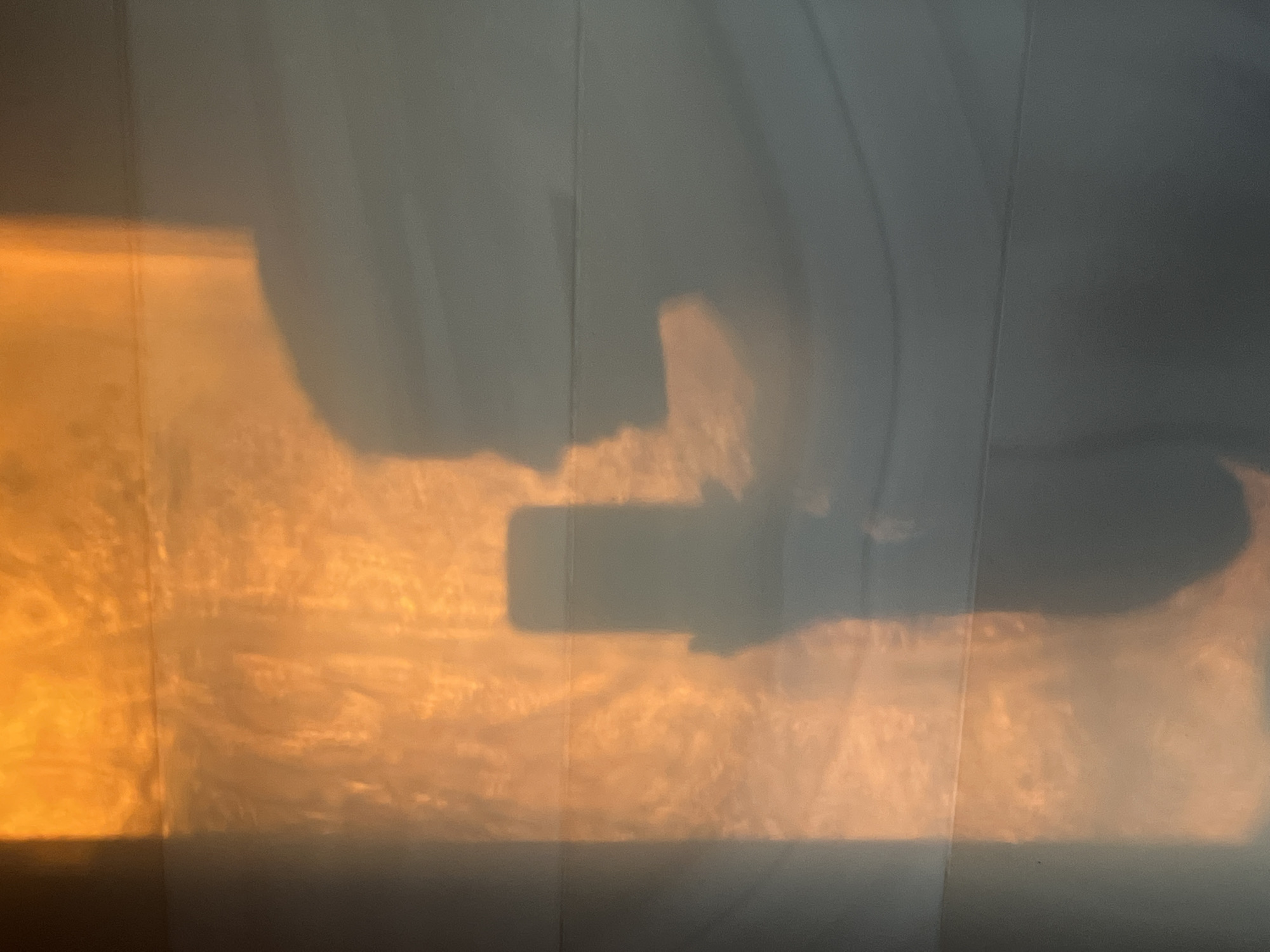 A diffused light with warm, orange hues illuminates a wall, creating shadows of objects placed in front of the light source. The shadows reveal the presence of a hand holding a rectangular object, potentially a phone, and draped material, possibly a curtain, is casting curved shadows on the wall. It is not possible to pinpoint the specific location within Riga, Latvia, based on the image alone.