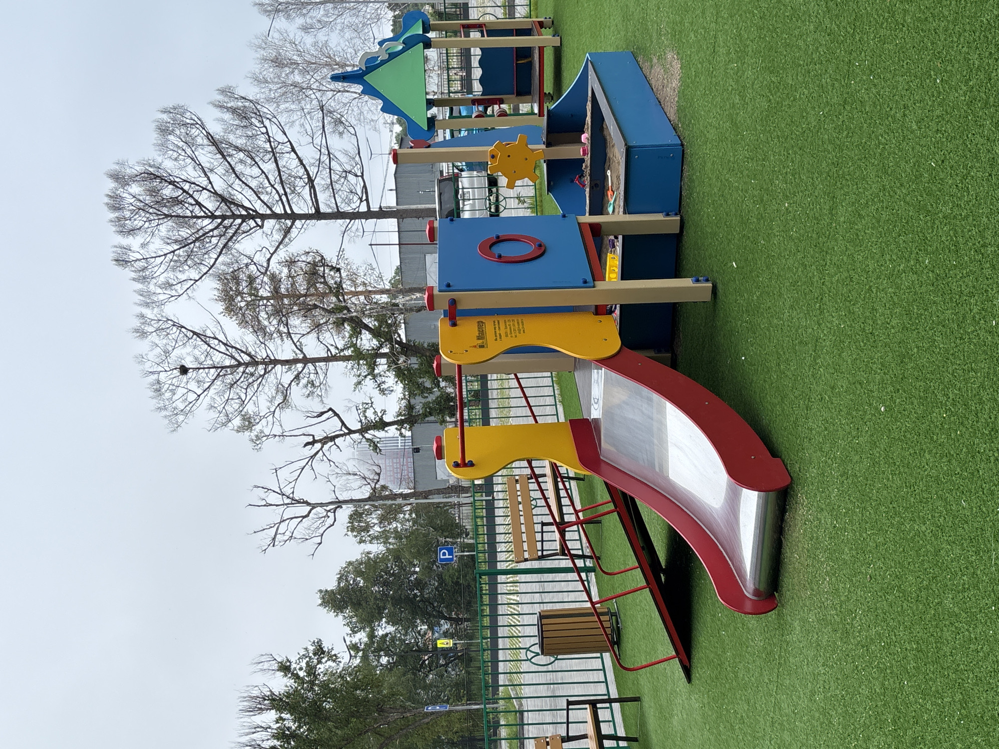 An outdoor children's playground is depicted on a cloudy day, likely in Blagoveshchensk, Russia. The playground is situated on a bright green artificial turf surface. The main play structure features a red and yellow slide with a silver chute, an accompanying ramp, and a blue sandbox with a yellow gear-like toy. Adjacent to this structure, a smaller blue and green gazebo-like playhouse is visible. In the background, a green metal fence encloses a paved area with parking spaces, and beyond that, trees with bare branches and some foliage are present, along with industrial-looking buildings. A trash can and two wooden benches are also part of the playground area. Several parking signs, including a blue "P" sign, are visible near the fence. No people are present in the image.