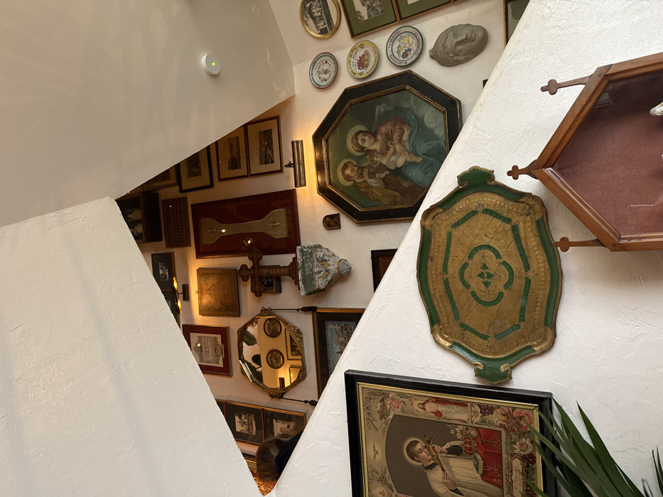 An interior scene located within the city of Barcelona, Spain, features a densely decorated wall, likely part of a staircase or an inclined architectural surface. The walls are white and textured. A prominent diagonal white beam or ceiling structure bisects the upper part of the frame.
The wall is completely covered with an eclectic collection of framed art, mirrors, and sculptural objects. Key items include:
* A large, centrally placed octagonal painting depicting the Holy Family (Mary, Joseph, and a child Jesus), set within a dark frame.
* To the left and slightly below, a large rectangular framed print shows a male saint holding a crucifix to his chest, with Jesus in the background.
* Above and to the left of the Holy Family painting, an ornate hexagonal mirror with a gold frame reflects parts of the room.
* Below the mirror, a dark wooden crucifix is mounted on an elaborate, carved wall corbel with green and gold accents.
* Adjacent to the crucifix, a tall, narrow, white object with a central cross shape is mounted on a red background.
* Further to the right, a collection of decorative plates and a gray classical-style head bust are arranged on the wall.
* In the lower central area, a large, ornate, antique serving tray with a green border and a gold center featuring a quatrefoil design is mounted horizontally.
* In the bottom right corner, a small, wooden, house-shaped shrine or reliquary box with cross-shaped finials is visible.
* Numerous other framed items, including smaller landscapes, certificates, and religious images, fill the remaining wall space.
* Several small spotlights illuminate specific artworks.
* A white smoke detector with a green indicator light is visible on the upper right ceiling.
* The top of a person's head with dark hair is partially visible on the far left, positioned below the diagonal white structure.
* Green foliage from a plant is visible in the bottom left corner.