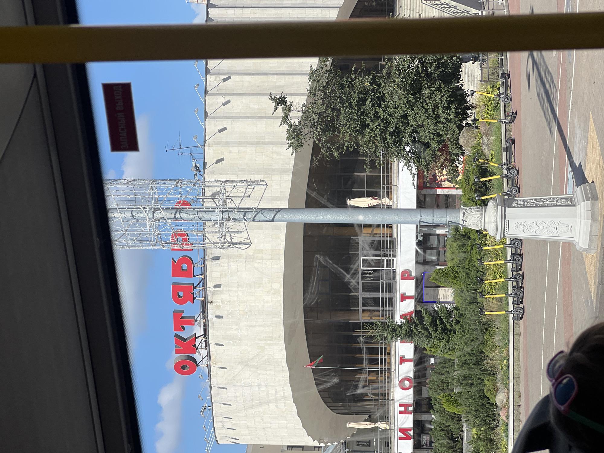 The image depicts the facade of a large building, likely a cinema or theater, with the prominent red Cyrillic lettering "ОКТЯБРЬ" (October) above the entrance. Below this, the word "КИНОТЕАТР" (Cinema) is also visible in red lettering. The building's architecture is characterized by a large, curved concrete facade with a row of windows. To the left of the entrance, a flag of Belarus is displayed. In front of the building, there is a row of parked electric scooters. The scene is viewed from inside a vehicle, possibly a bus, as indicated by the window frame and a yellow vertical pillar on the right. A small red sign with white Cyrillic text "ЗАПАСНЫЙ ВЫХОД" (Emergency Exit) is visible through the window on the upper right. The sky is blue with some clouds. The location is the "October" Cinema in Minsk, Belarus.