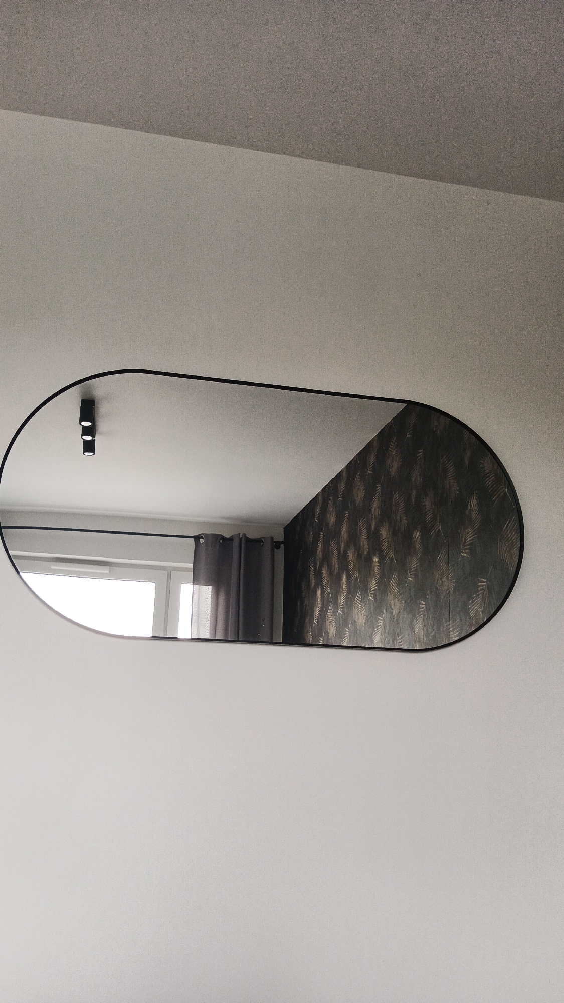 The image shows an oval mirror with a black frame mounted on a light-colored wall. The mirror reflects a room interior. In the reflection, a window with white frames and gray curtains is visible on the left. Above the window, a black track light with three cylindrical fixtures is attached to the ceiling. On the right side of the reflection, a section of dark wallpaper with a gold-leaf pattern is visible. The mirror's surface is clean, and the lighting appears to be ambient. The image was captured in Zielona Góra, Poland.
