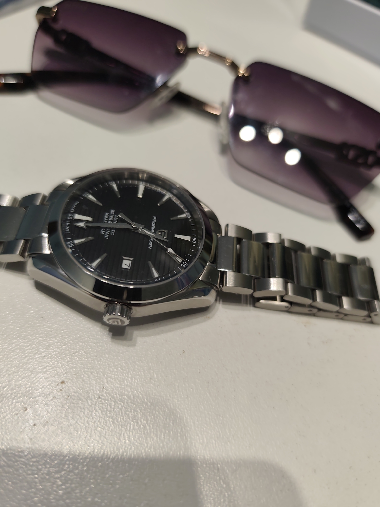 The image displays a metallic wristwatch and a pair of sunglasses resting on a light-colored, textured surface.
The wristwatch, positioned in the foreground, features a silver-colored stainless steel case and bracelet. Its black dial, marked "PAGANI DESIGN," displays "AUTOMATIC," "WATER RESISTANT 10BAR / 100M," and "JAPAN MOVT." White hour markers, including triangular ones at 12, 6, and 9 o'clock, contrast with the dial's subtle horizontal ridged pattern. Sword-shaped hour and minute hands, along with a thin second hand, are silver-toned. A date window at the 3 o'clock position shows the numeral '7'. The crown, located at 3 o'clock, bears a circular logo. The watch's bracelet is a three-link oyster-style design with both brushed and polished finishes.
Behind the watch, a pair of rimless sunglasses with purple-tinted lenses and dark-patterned arms is partially visible and out of focus. Reflections of circular light sources are present on the sunglass lenses.
The surface beneath the objects is off-white and displays a fine, irregular texture, along with minor dark specks. Illumination appears to be from above, creating subtle shadows beneath the watch and a diffused shadow extending from the blurred sunglasses. The background is out of focus, with an indistinct light-colored object visible in the upper right. The specific location in Pazardzhik, Bulgaria, cannot be determined from the visual information provided in the image.