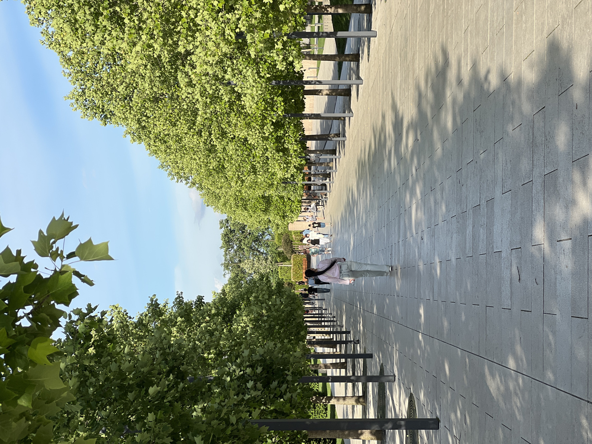 The image shows a long, tree-lined pedestrian walkway in Krasnodar, Russia. The walkway is paved with rectangular gray stones and is bordered on both sides by rows of trees with green foliage, supported by dark metal structures. Shadows from the trees fall across the walkway. Several people are walking along the path, including one figure in the foreground wearing light-colored clothing and carrying a bag. The sky is blue with some light clouds visible above the trees.
