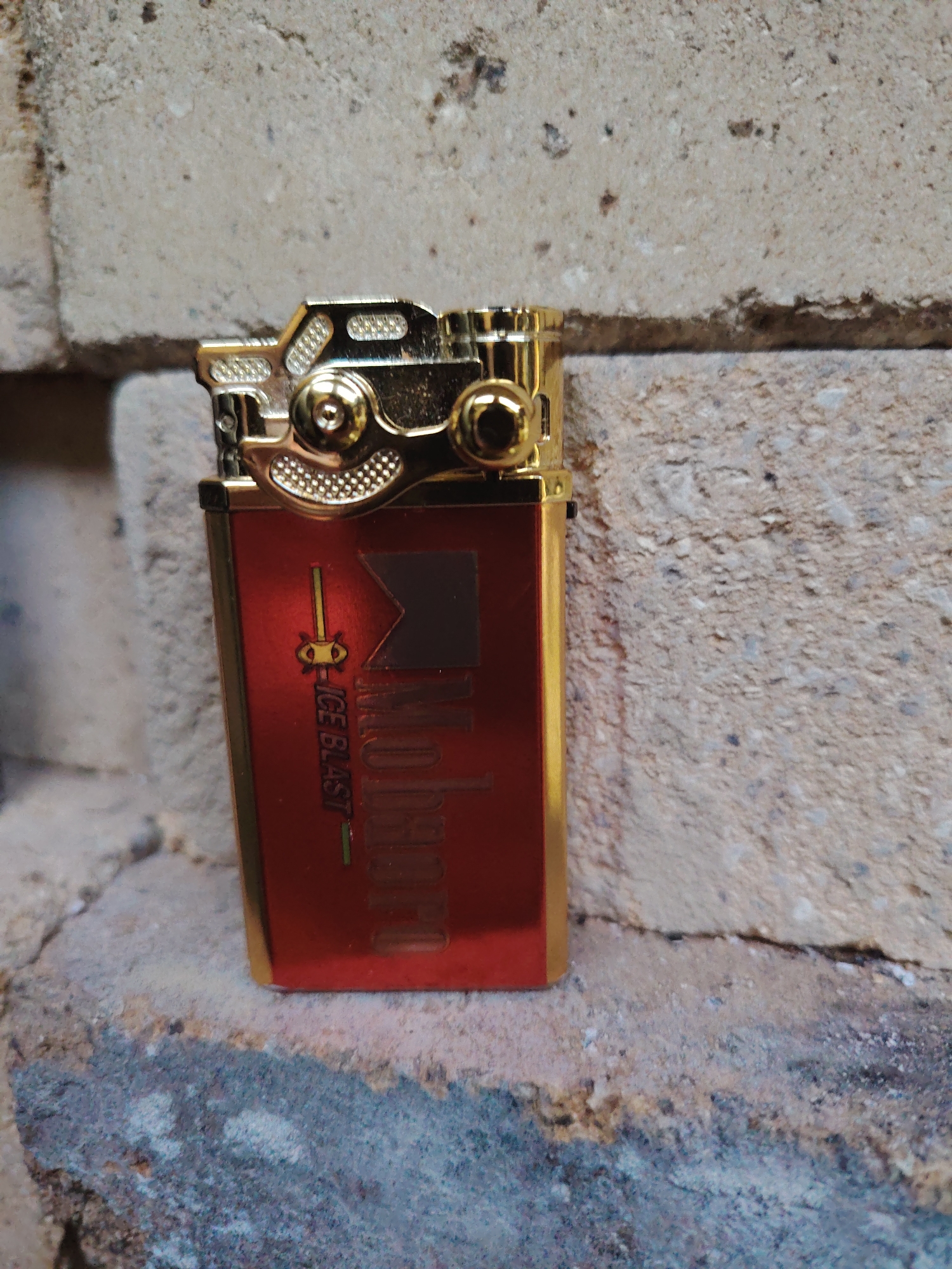 A gold and red lighter with the Marlboro logo and the text "ICE BLAST" is positioned on a rough, grey brick surface. The lighter is upright and slightly angled to the left. The background consists of more of the rough, grey brick wall. The scene appears to be outdoors, with natural light casting shadows. No people are present in the image. The location is described as Malakhovka, Russia.