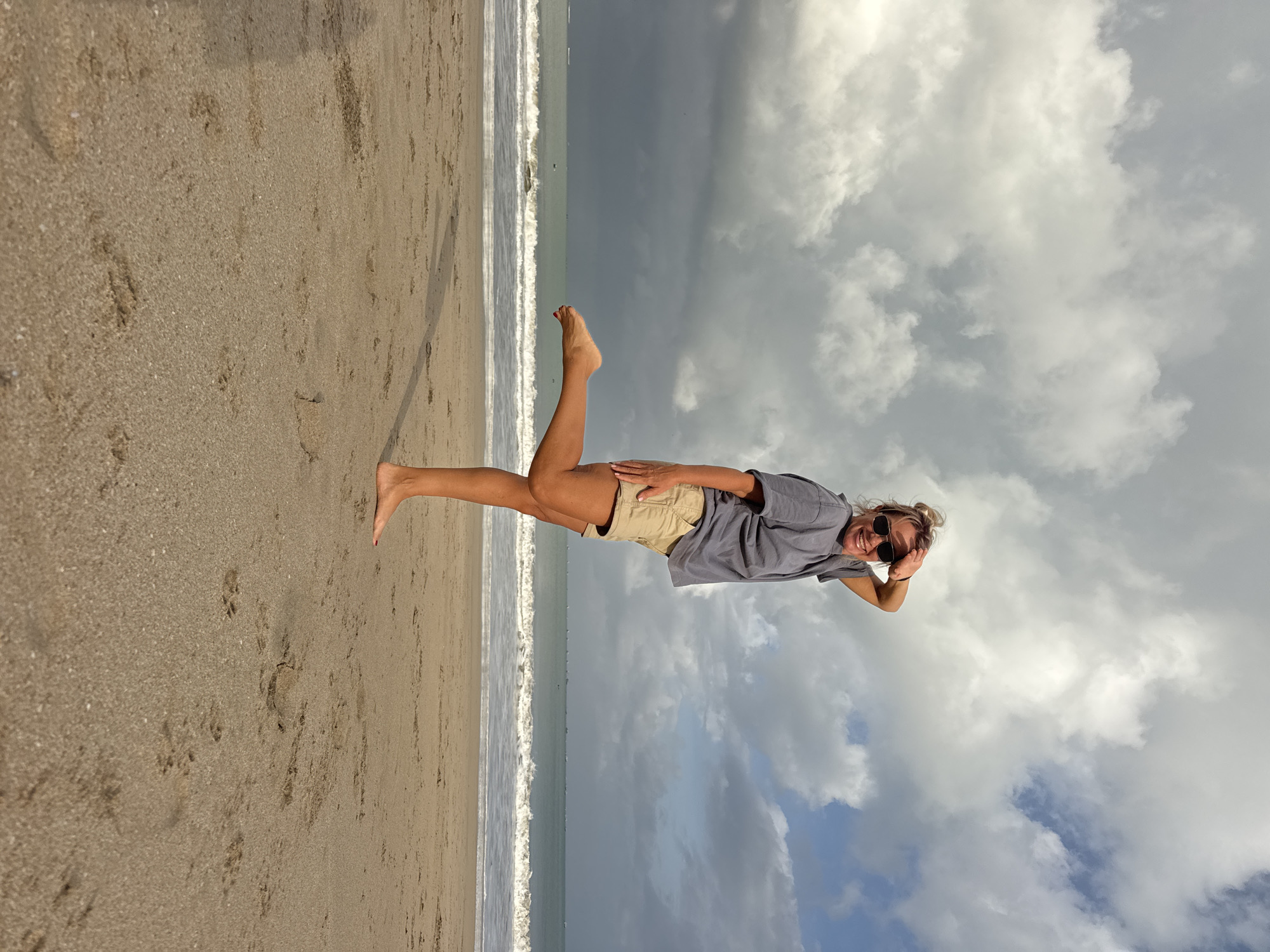 A woman with blonde hair pulled up in a bun and sunglasses stands on a sandy beach in Kuta, Indonesia, with her left leg bent and raised, and her right hand on her forehead as if shielding her eyes from the sun. She is wearing a gray t-shirt and tan shorts. The ocean is behind her, with waves breaking on the shore. The sky is filled with dramatic clouds, with patches of blue sky visible. The sand is a light brown color, and there are footprints visible in the sand. The overall scene is one of a casual beach outing.