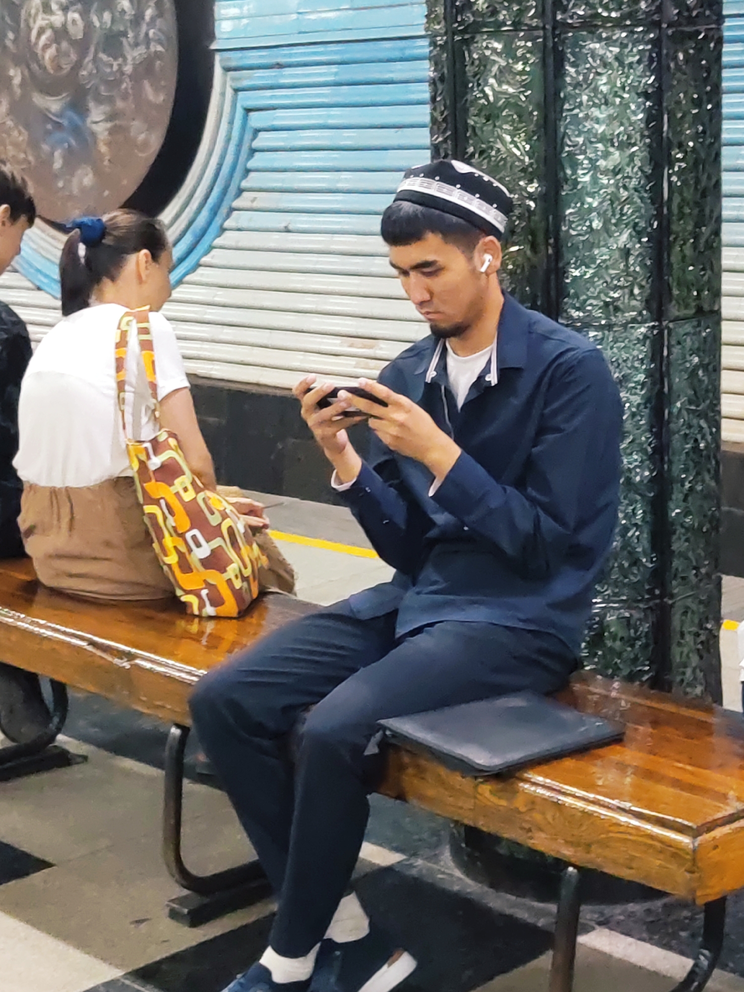 A young man wearing a black and white kufi and a dark blue shirt with a white undershirt, is seated on a wooden bench and looking down at his mobile phone. He is wearing earbuds and has a beard. He is sitting next to a woman with a ponytail, wearing a white t-shirt and brown pants, who is carrying a patterned bag. The bench is in front of a wall with a blue and white striped pattern and a tall, dark green, textured pillar. The floor is tiled in a black and white checkered pattern. This scene appears to be from a metro station in Tashkent, Uzbekistan, given the architectural style and common attire.