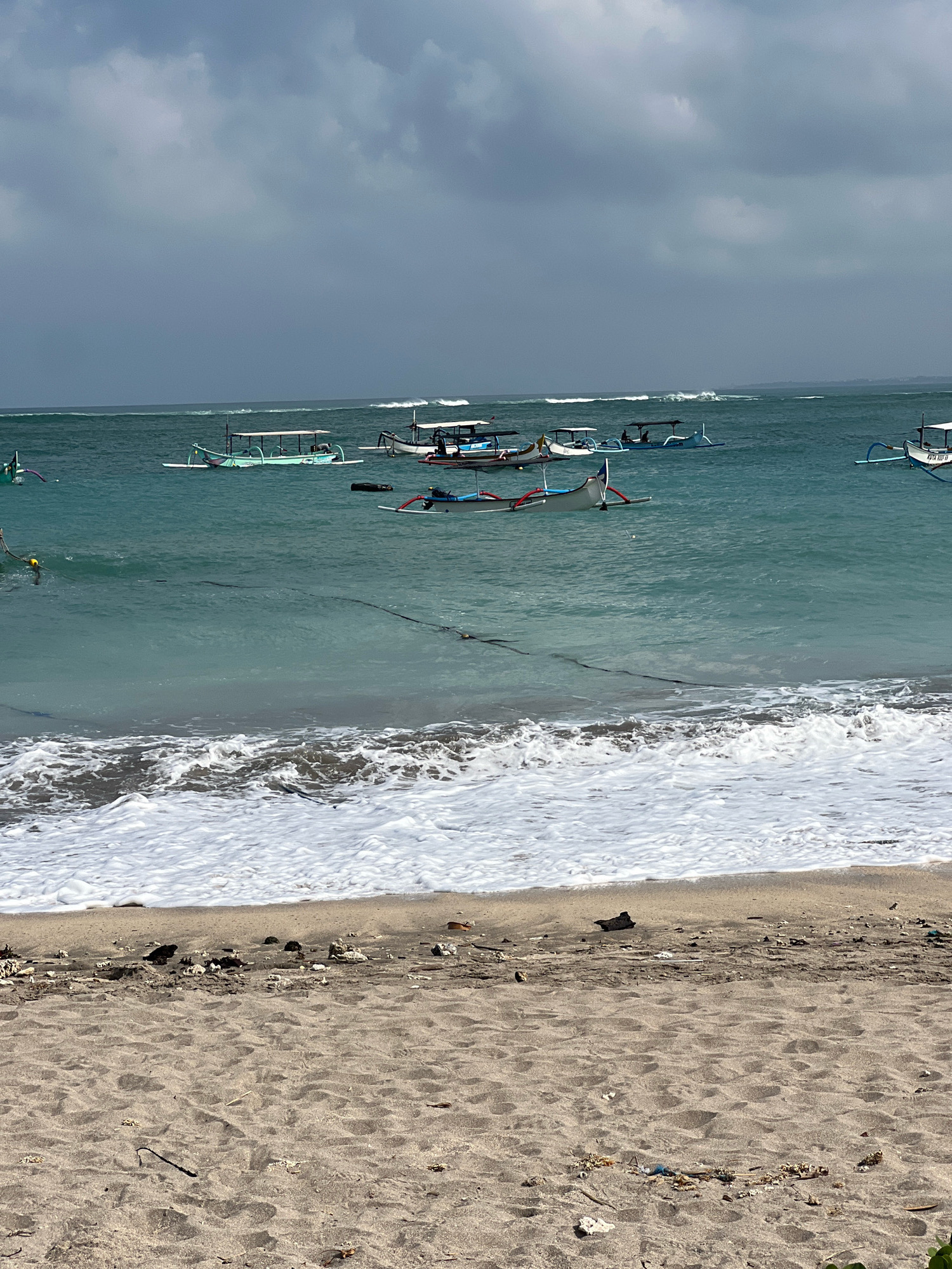 The image captures a view of the ocean and beach in Kuta, Indonesia. Several traditional Balinese fishing boats, known as jukung, are visible on the turquoise water. Some of these boats are moored, while others appear to be actively used. The water is relatively calm with gentle waves breaking on the shore, creating a foamy white surf. The sky is overcast with grey clouds, suggesting a potentially stormy or humid weather condition. The beach in the foreground is sandy, with some debris and small pieces of coral scattered on its surface. The scene depicts a typical coastal environment in Kuta, known for its beaches and fishing activities.