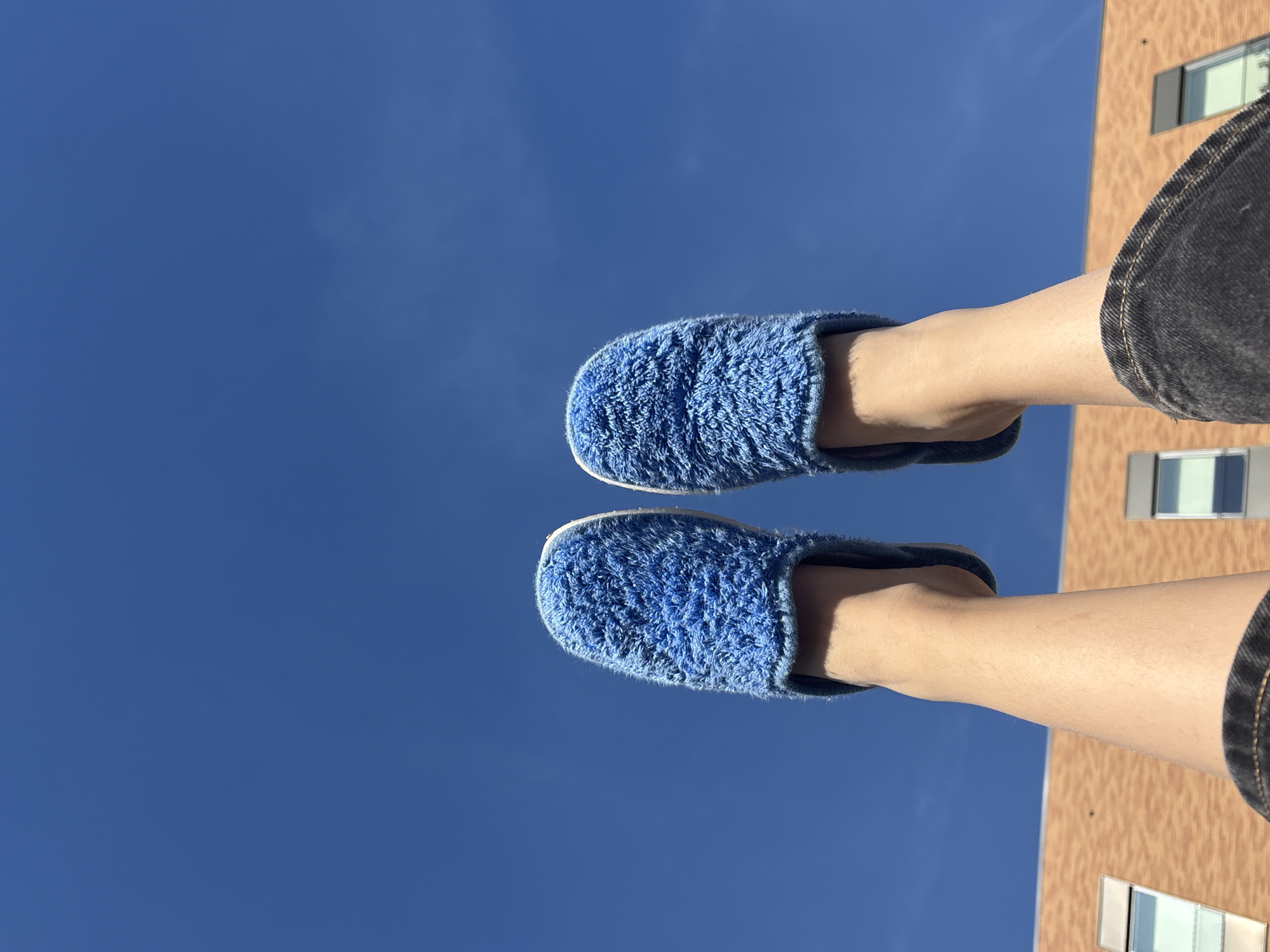 The image depicts a person's legs and feet wearing blue, fuzzy slippers against a backdrop of a clear, bright blue sky. A portion of a tan-colored building with evenly spaced windows is visible along the right edge of the frame. The person is wearing dark-colored jeans or shorts. The photo appears to be taken from a low angle, looking up at the person's feet against the sky and building. There is no information to suggest the location of the photo is in Milan, Italy.