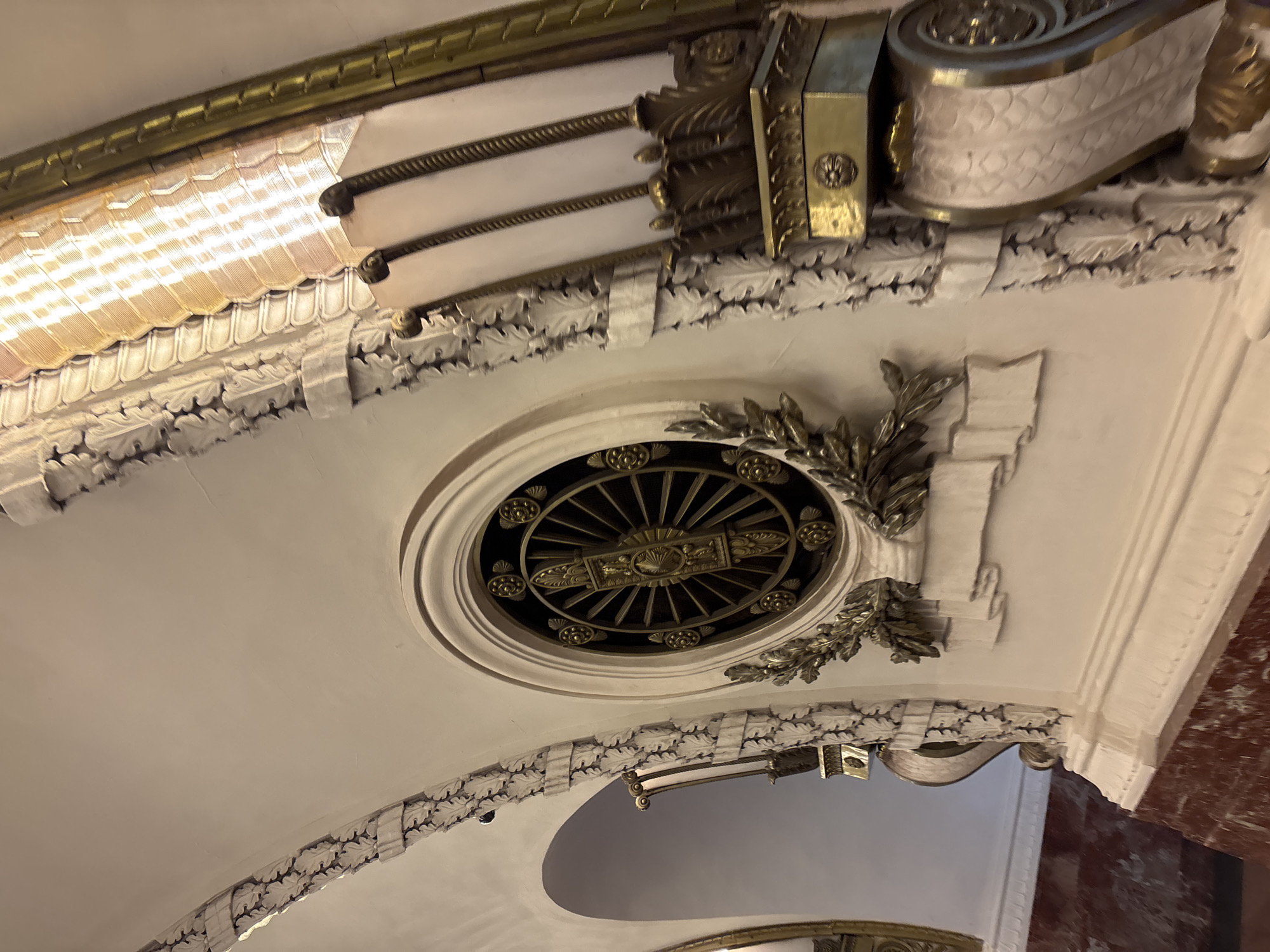 The image depicts an ornate ceiling detail in Saint Petersburg, Russia. The ceiling is white with gilded accents, including a circular vent with a radial design. The vent is flanked by decorative leaf motifs and sculpted architectural elements. A bright, diffused light fixture is visible along the upper left edge. The lower portion of the image shows a curved archway and the top of a golden structure, suggesting the location is within a grand interior space. A reddish-brown marble or stone wall is visible to the right.