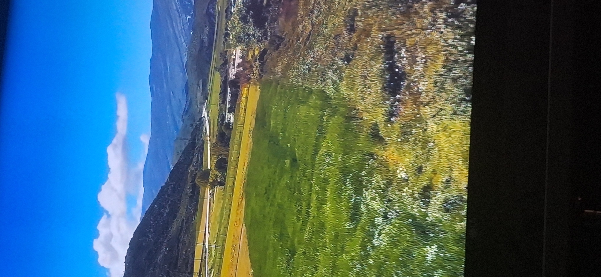 The image shows a scenic view of rolling hills and fields under a bright blue sky with scattered clouds. The foreground is dominated by a lush green meadow, interspersed with patches of yellow and white wildflowers. In the midground, there are fields of yellow and green crops, separated by a fence. Further back, a winding road or river can be seen, bordered by trees. Beyond that, the landscape transitions into darker, undulating hills and mountains in the distance. The scene is bathed in natural sunlight, suggesting an outdoor setting in a rural or mountainous area. The image does not contain any elements that indicate it is located in Bila Tserkva, Ukraine.
