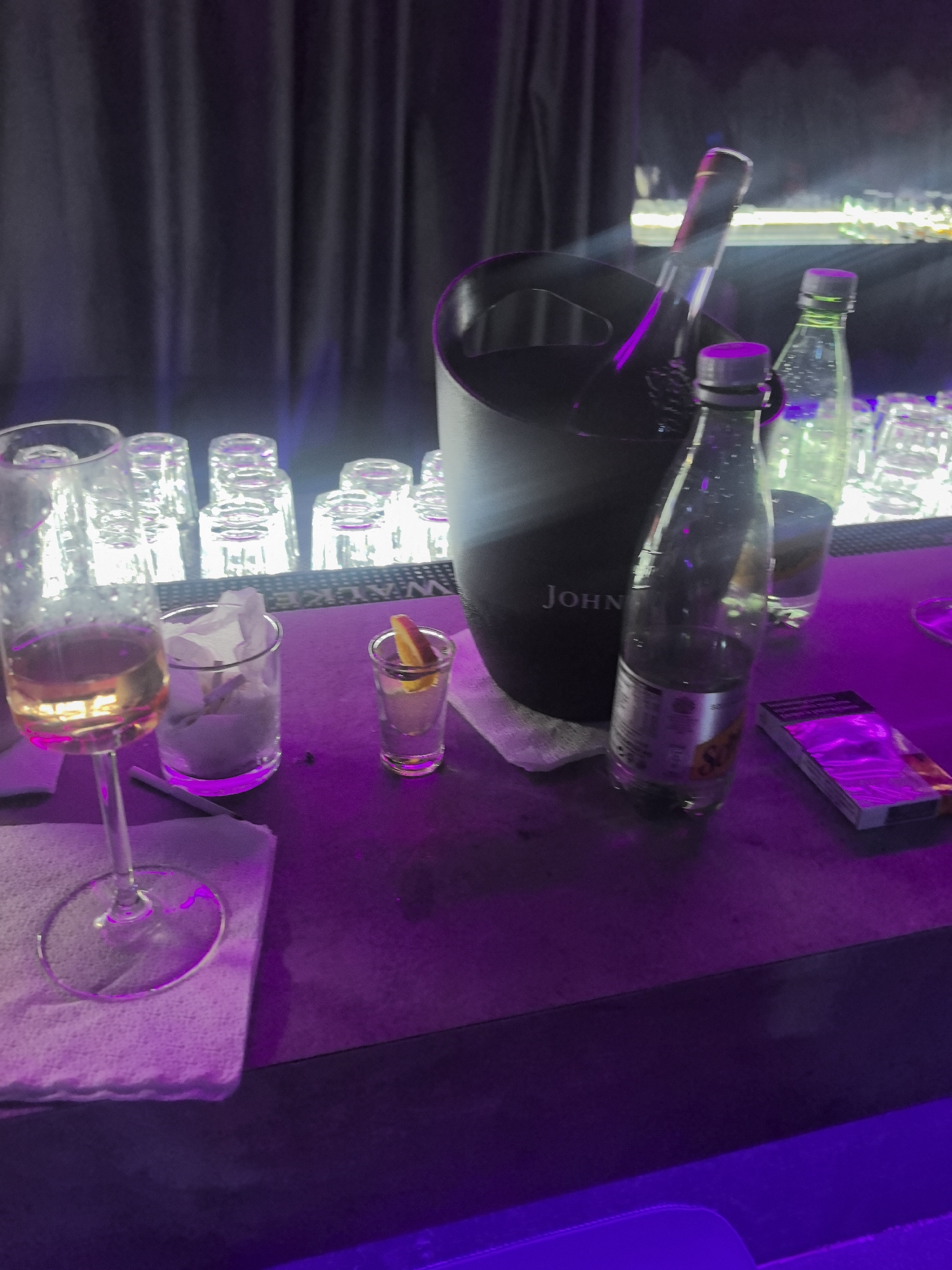 The image shows a dimly lit bar scene in Stara Zagora, Bulgaria, featuring an ice bucket labeled "John," holding a bottle of champagne. To the left, there is a wine glass filled with liquid, a small glass with a citrus garnish, and a glass containing tissues. Two sealed bottles of liquid are present near the ice bucket, and a packet of cigarettes is placed on the surface. In the background, clean empty glasses are arranged on a shelf. The environment is a dark, possibly club-like setting with purple lighting.