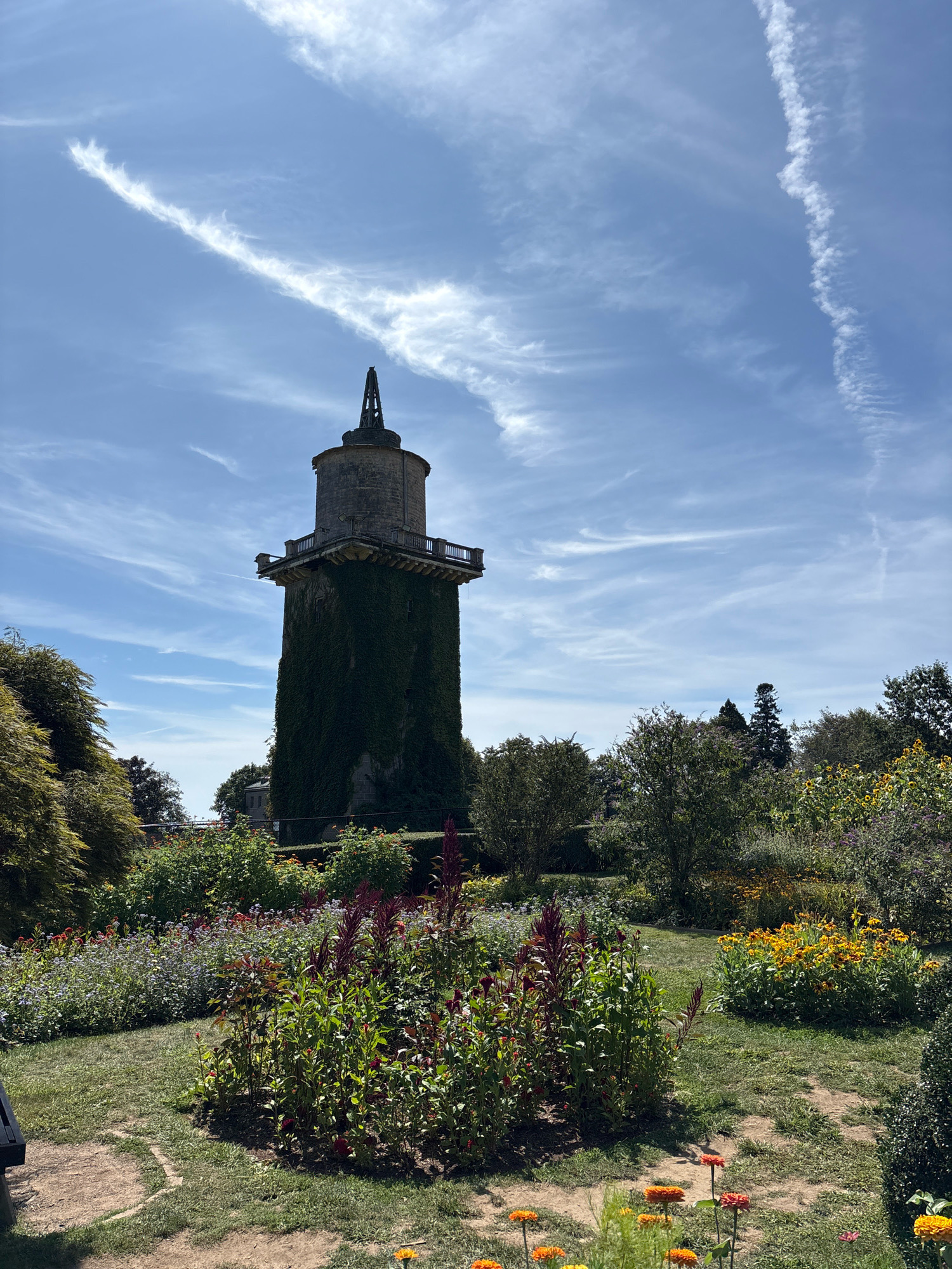 This image depicts a picturesque garden with a prominent, ivy-covered tower in the background. The tower, made of stone, has a balcony-like structure near its top and a pointed spire. It appears to be an old structure, possibly a water tower or a folly, integrated into the landscape.
The foreground is filled with a vibrant flower garden, showcasing a variety of colorful blooms, including yellow flowers, red and purple plants, and smaller white and pink blossoms. The garden is well-maintained with manicured grass paths winding through the flowerbeds. Lush green trees and shrubs surround the garden and the tower, creating a serene and natural setting.
The sky above is a clear blue with scattered wispy white clouds, suggesting fair weather. The overall scene is one of natural beauty and tranquility, a peaceful retreat within a lush garden environment. It is not possible to determine the specific location in Waterford, United States of America, from the image alone.