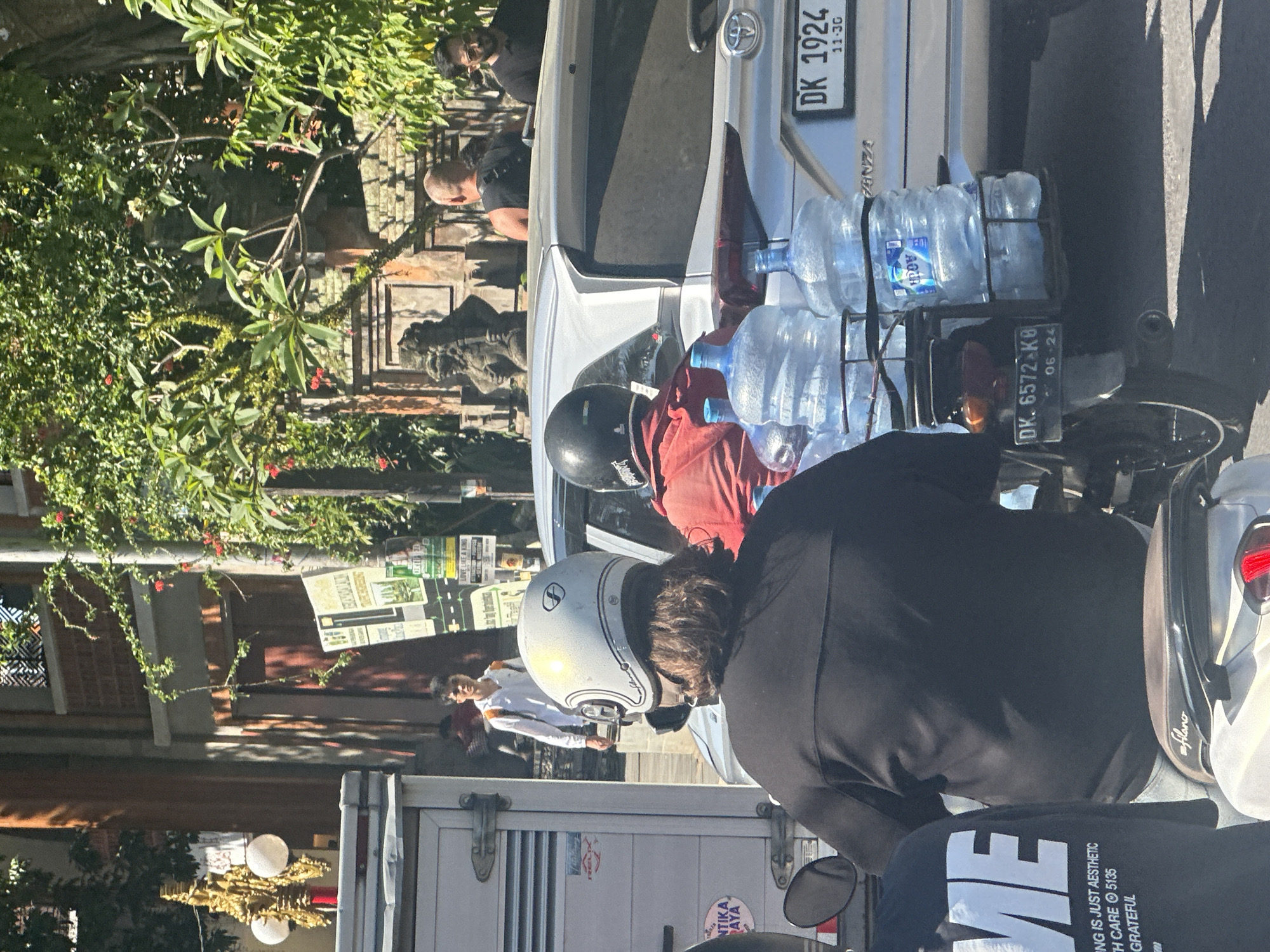 In Ubud District, Indonesia, on a sunny day, a person wearing a black jacket and a white helmet rides a scooter carrying a rack of six large water bottles. Behind them, a person in a red shirt also rides a scooter. To the right, a silver car with a license plate reading "DK 1924" is visible. In the background, a man in a white shirt walks past a truck and a building with traditional Balinese architecture. Lush green trees and foliage are present throughout the scene.
