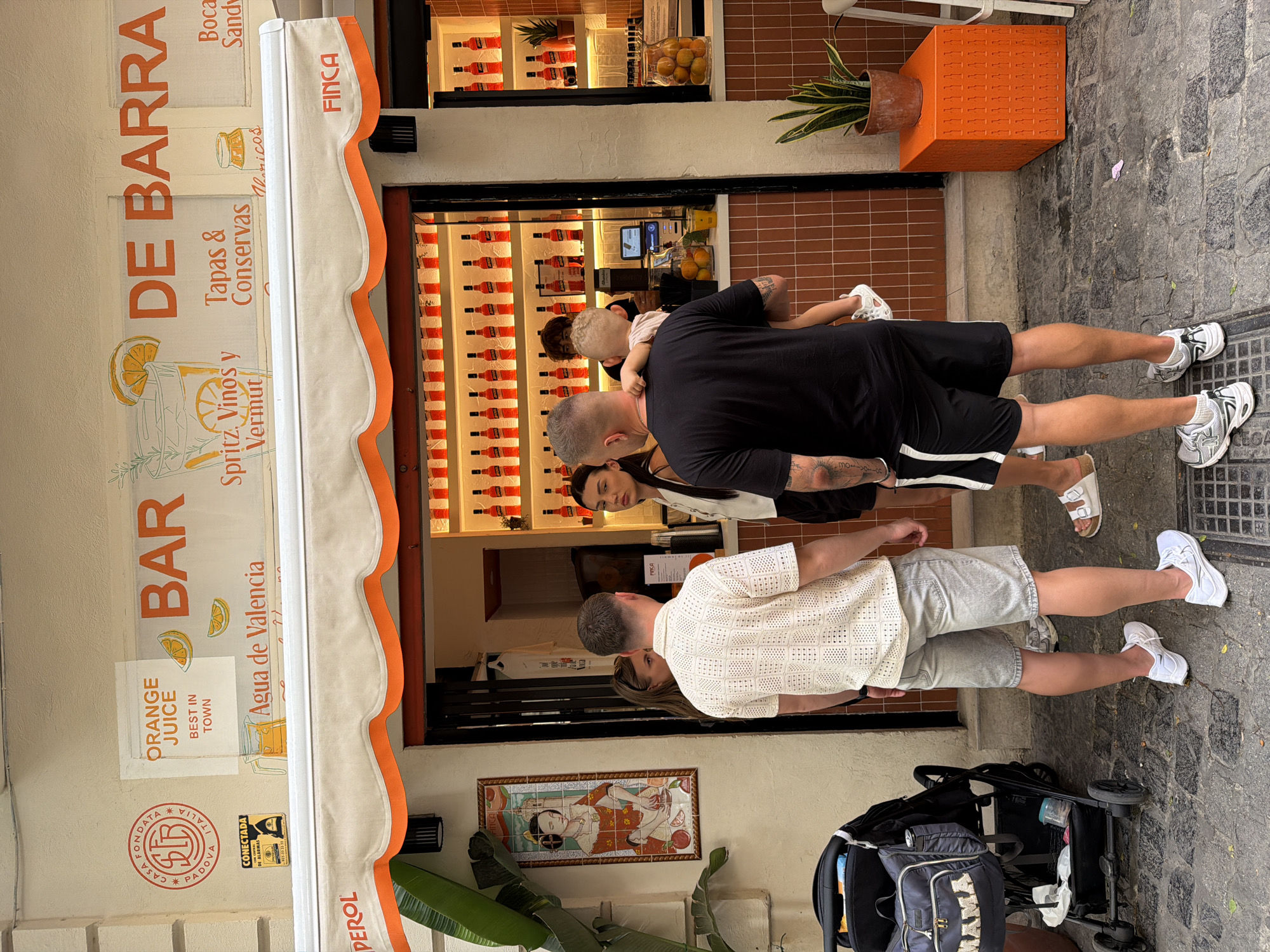 In an outdoor setting, possibly a street or alleyway, a bar with an orange and white awning is depicted. The awning has "FINCA" printed on it. Above the awning, a sign reads "BAR DE BARRA". Various signs with text such as "ORANGE JUICE BEST IN TOWN" and "Agua de Valencia" are visible. A few people are gathered outside the bar, including a man holding a young child, a woman in a white top, and a man in a patterned shirt and shorts. A baby stroller is parked to the left of the scene. The bar interior is visible, showing shelves stocked with numerous red bottles. An orange cube-shaped object and a potted plant are situated on the ground to the right. The ground is paved with cobblestones.