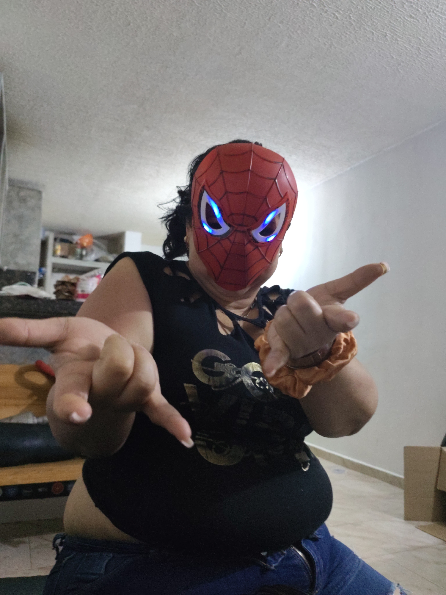 The image shows a person wearing a red Spider-Man mask with glowing blue eyes. The person is wearing a black t-shirt with a graphic print and blue ripped jeans. The person is holding up their hands in front of them, with one hand extended towards the camera. An orange scrunchie is visible on the person's wrist. The background appears to be an indoor setting with a ceiling light, a counter with items on it, and a wooden bench or furniture. The scene is consistent with a domestic environment. The image does not contain any specific visual cues that would place it in the city of Piloncito, Venezuela.
