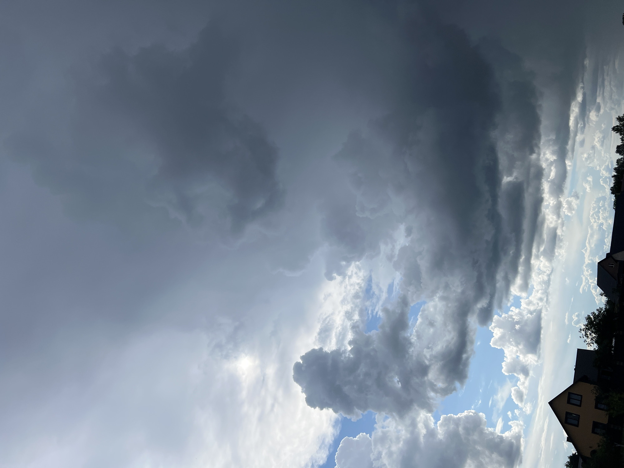 The image captures a dramatic sky over Sobishchytsi, Ukraine, characterized by a stark contrast between bright, sunlit clouds and dark, ominous storm clouds. The foreground features the rooftops of several houses, silhouetted against the lighter parts of the sky. The overall impression is one of impending weather change, with the turbulent cloud formations dominating the upper two-thirds of the frame.
