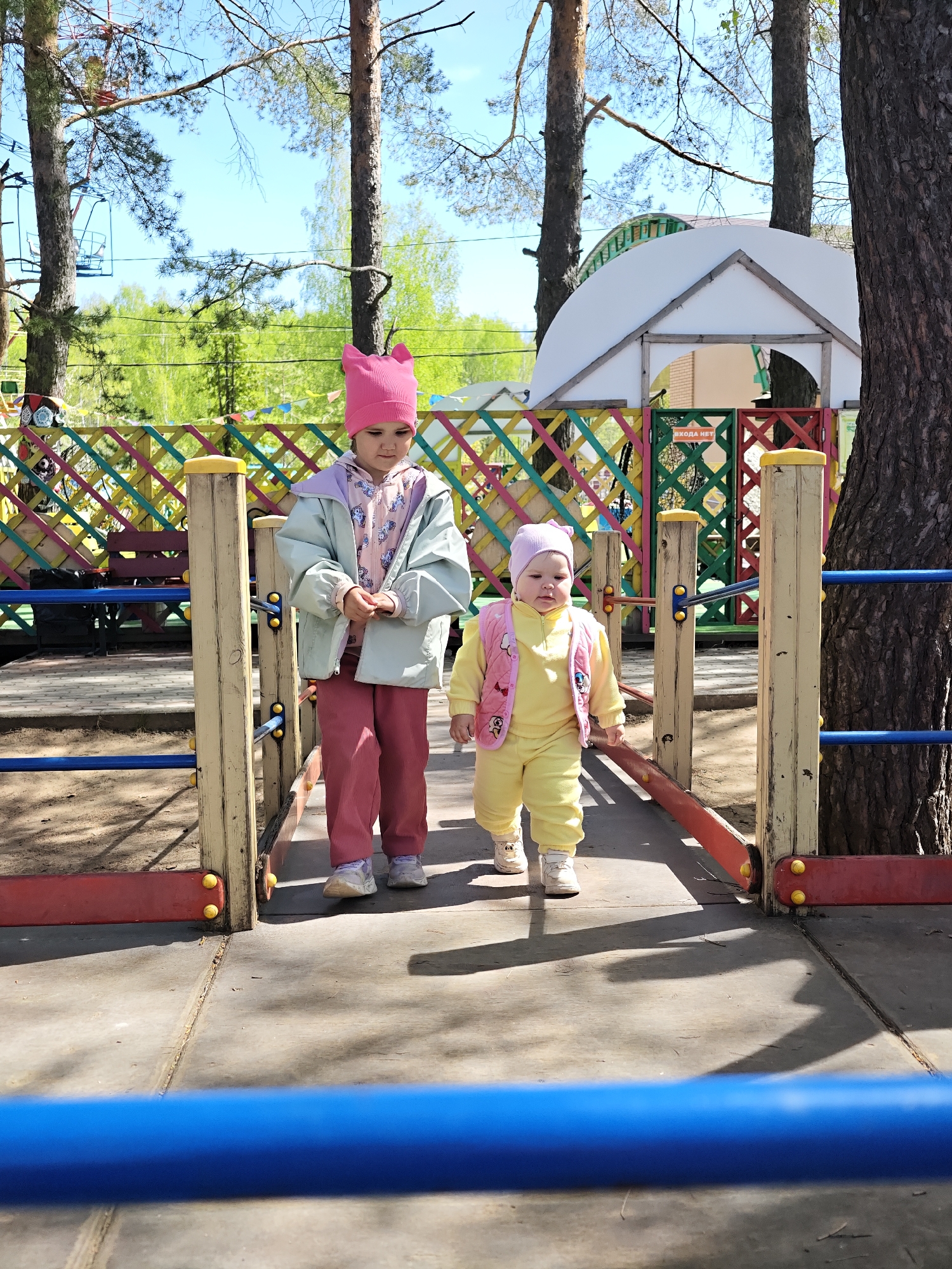 The image shows two young children, likely sisters, walking on a playground structure in what appears to be a park. The older child, on the left, is wearing a pink cat-eared hat, a light green jacket, a pink hooded shirt, and pink pants. The younger child, on the right, is wearing a yellow jumpsuit and a pink vest with a matching hat. They are on a bridge-like structure with wooden posts and colorful railings. In the background, there's a colorful fence, trees, and a white dome-shaped structure. Based on the location information, this is in the city of Chaikovsky, Russia.