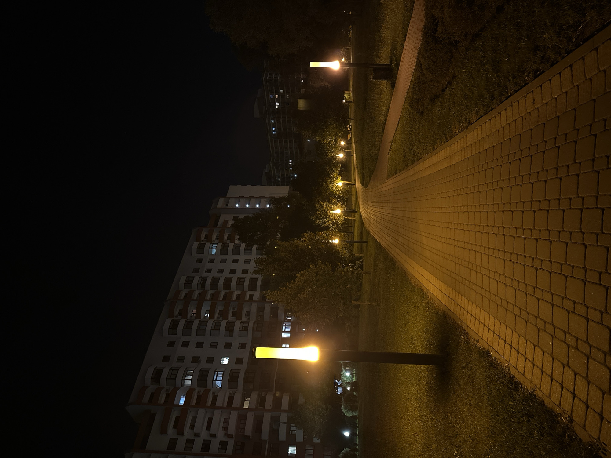 The image depicts a nighttime view of a park path illuminated by lampposts. A wide, paved walkway, constructed of rectangular bricks, stretches into the distance, curving slightly to the left. On either side of the path, grass lawns are visible, with several trees scattered throughout. In the background, a large, multi-story apartment building with visible lit windows stands to the left, and another apartment building can be seen further in the distance to the right. The sky is dark, indicating it is nighttime. Several lampposts with a warm, yellow glow are placed along the path, casting light on the surrounding area. One prominent lamppost is in the foreground on the left, and others are spaced out along the walkway. The scene is quiet and appears to be a residential area.