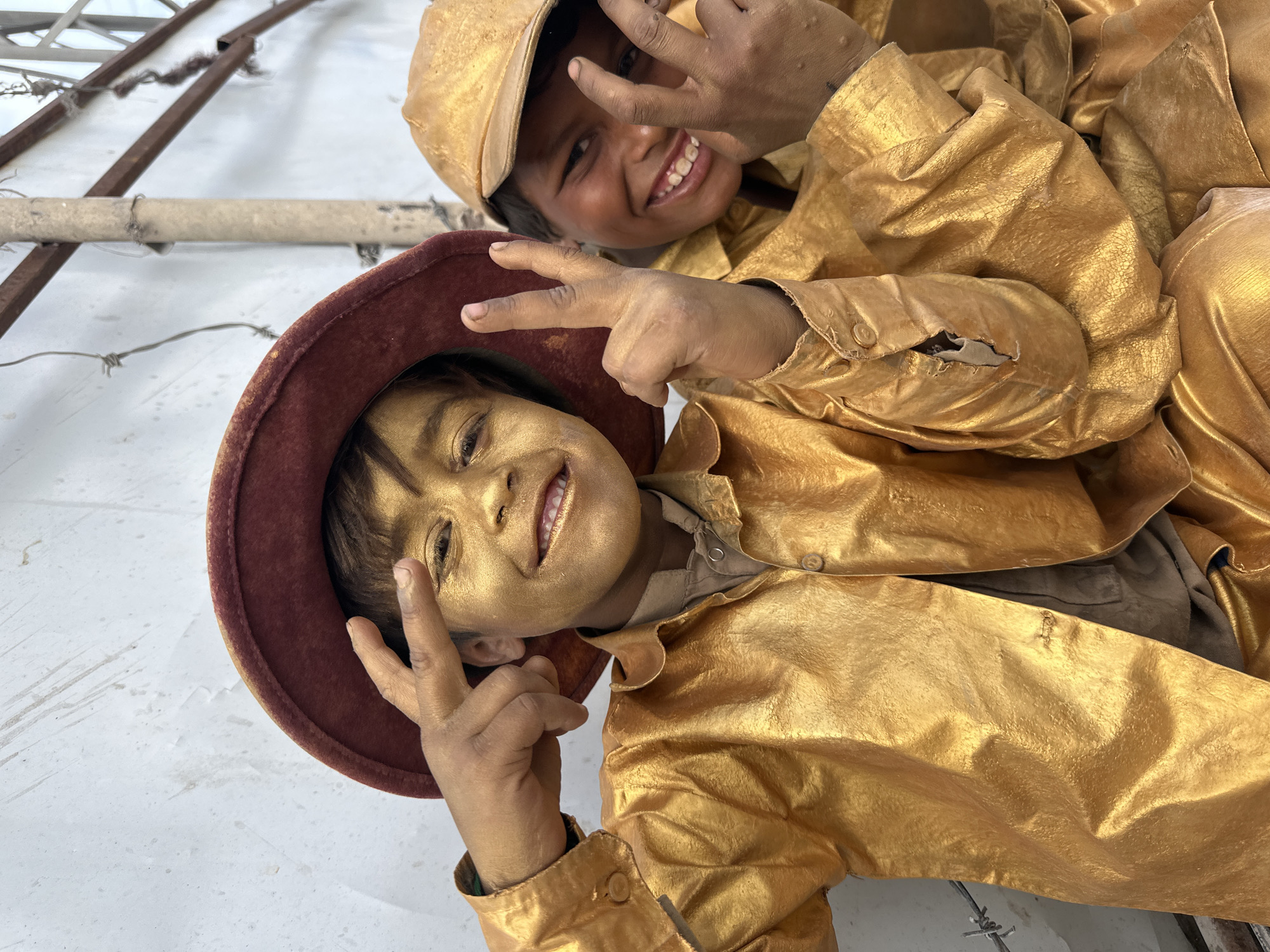 Two children covered in gold paint pose for a photo outdoors. One wears a burgundy hat, while the other sports a gold cap. They are both making the peace sign. The setting appears to be Quetta City Tehsil, Pakistan, based on the presence of exposed rebar and a rough, unfinished wall.