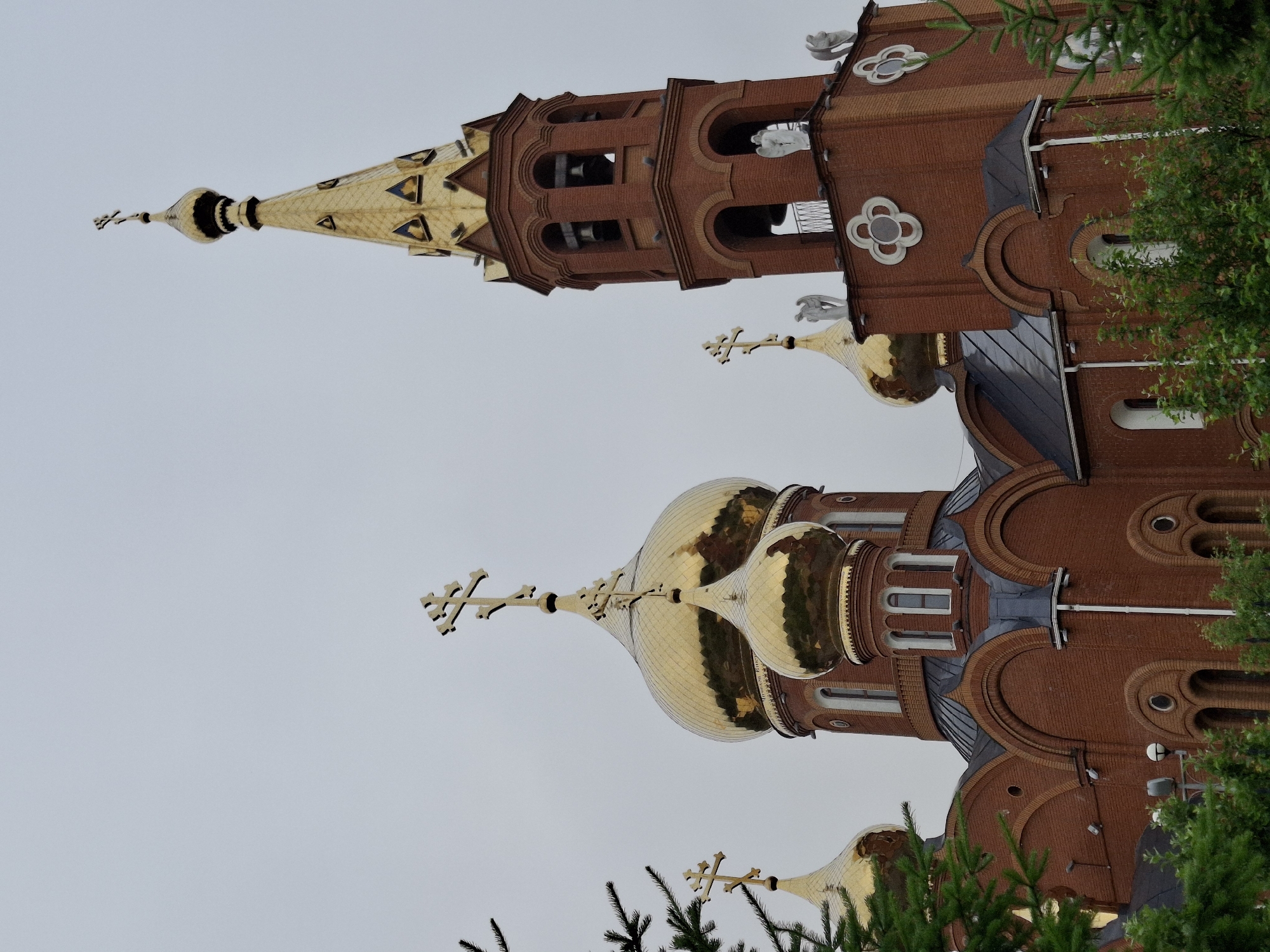 An Orthodox church with red brick construction and golden domes stands under a cloudy sky. The church features multiple onion-shaped domes, each topped with a cross, and a tall, slender bell tower also crowned with a golden, cross-topped spire. Sculpted angelic figures are positioned on the upper levels of the bell tower. The lower parts of the building are partially obscured by green evergreen trees and foliage. This is the Holy Trinity Cathedral in Chaikovsky, Russia.