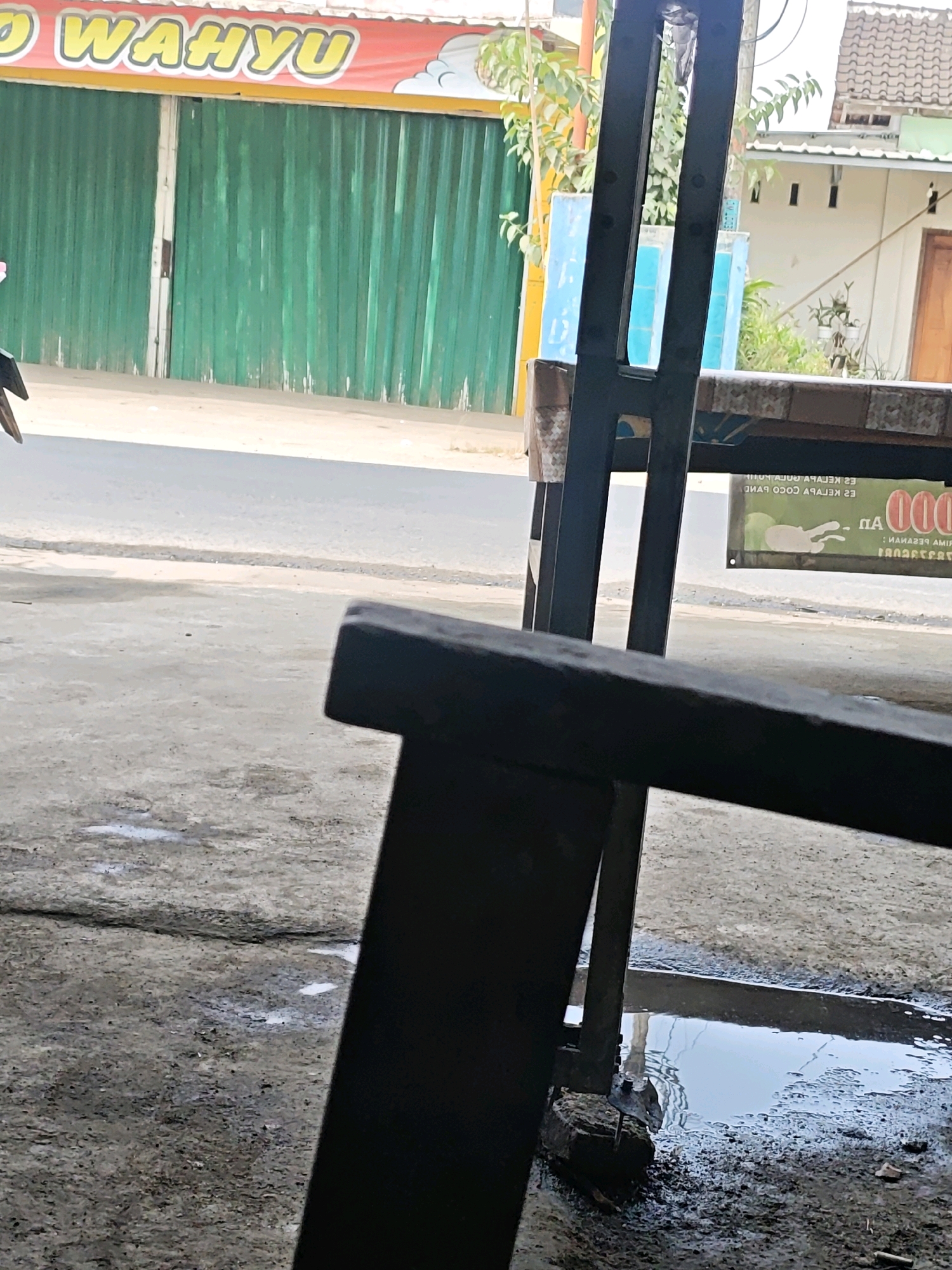The image depicts a street scene in Kemuning, Indonesia. In the foreground, a dark metal bench is positioned on a wet, concrete surface. Beyond the bench, a road is visible, with a row of green, corrugated metal shutters forming the facade of a building on the left. Above the shutters, a red banner with yellow lettering reads "WAHYU". To the right of the road, a second building with a tiled roof and a brown door is partially visible. A sign, seemingly advertising a food or drink establishment, is also present on the right, featuring green and white text. The overall environment appears to be an outdoor commercial area, possibly a shop or restaurant.