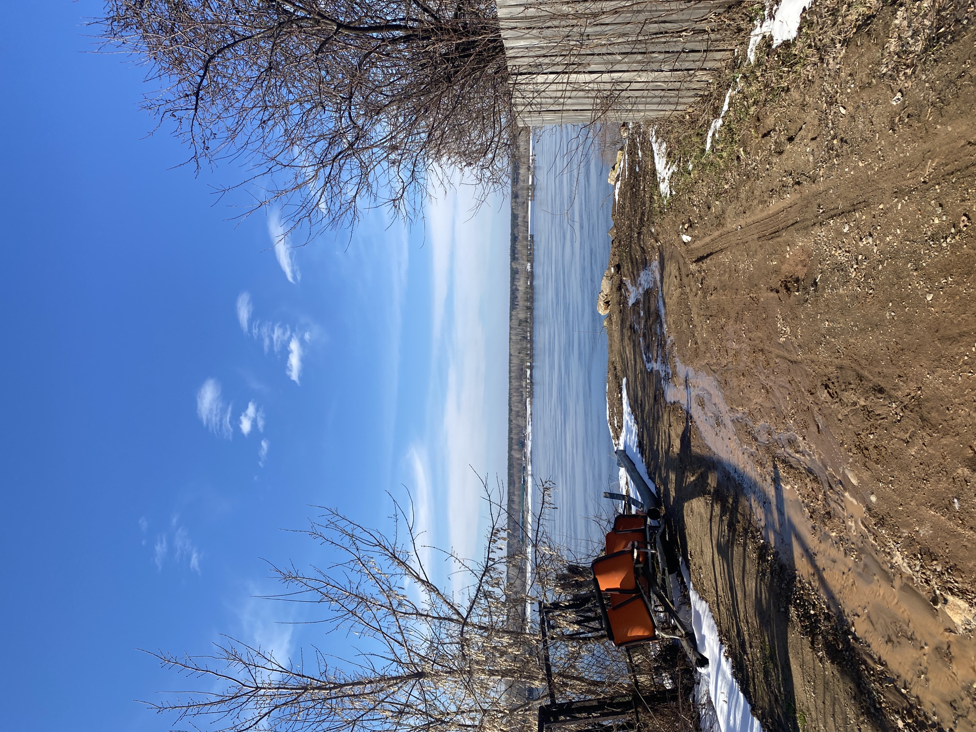 The image depicts a riverbank in Ольховка, Russia. The view is predominantly of a partially frozen river, with a clear blue sky overhead dotted with wispy clouds. Bare trees frame the top and bottom edges of the image. A muddy path runs along the riverbank, with patches of snow. Adjacent to the path is a stack of orange chairs. A wooden fence lines the top right edge of the frame. No people are visible. The scene suggests early spring or late winter.