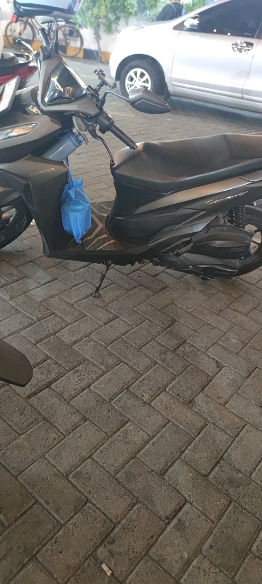 The image shows a close-up view of a dark-colored Honda scooter parked on a herringbone-patterned brick pavement. A blue plastic bag is hanging from the left side of the scooter. In the background, a white car is parked, and a bicycle is visible to the left of the car. The scene appears to be in an outdoor urban setting, likely Keboansikep, Indonesia, given the context.