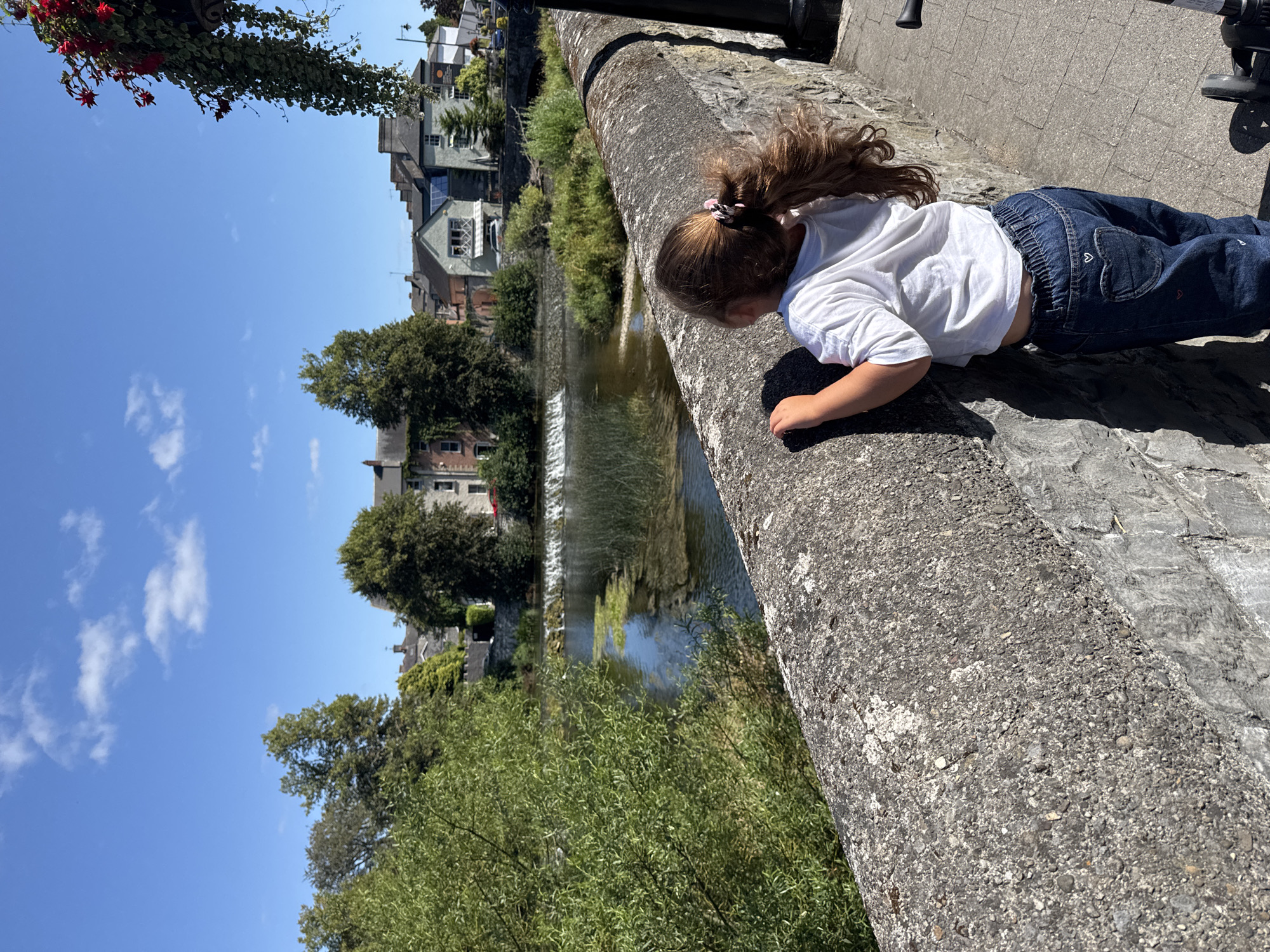 In Cahir, Ireland, a young girl with long, wavy brown hair tied back in a ponytail stands facing away from the camera, looking out over a river. She is wearing a white t-shirt and dark blue jeans adorned with a heart pocket detail. The girl is leaning against a thick, textured stone wall that runs along the foreground of the image. Beyond the wall, a river flows, with a small waterfall or weir visible in the mid-ground. The riverbanks are lined with lush green foliage and trees. In the background, a picturesque scene of stone buildings, likely houses and shops, with varied rooflines, is visible under a clear blue sky dotted with white clouds. Hanging plants with red flowers are visible in the upper right corner of the frame, suggesting a decorative element. The scene is bathed in bright sunlight, casting shadows on the wall and the pavement.