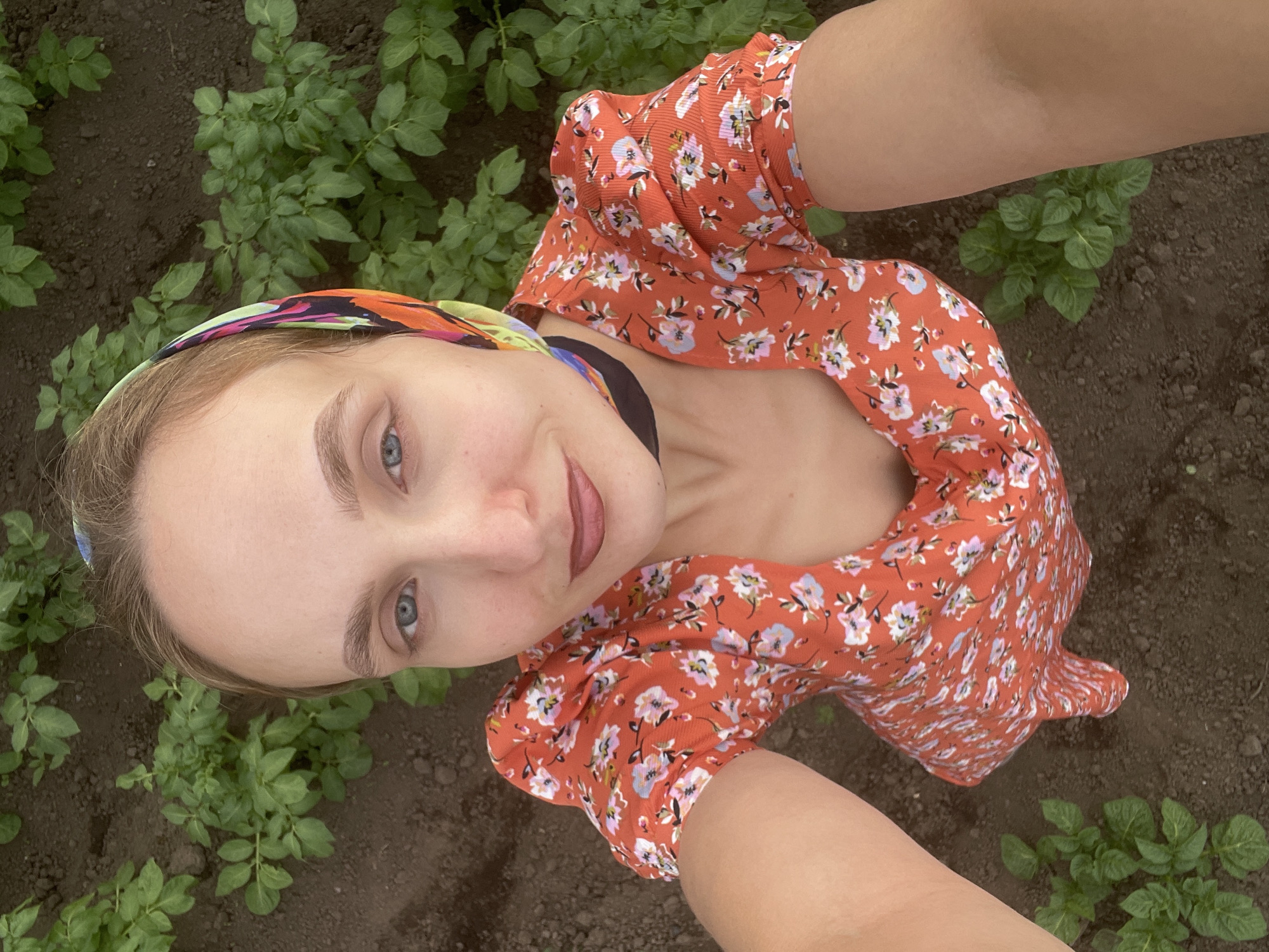 A high-angle, medium shot shows a woman with blonde hair and blue eyes taking a selfie. She is wearing a red dress with a floral pattern and a colorful headscarf. The woman is standing in a field of green plants, which appear to be potato plants, with dark soil between them. The background suggests an outdoor agricultural setting, possibly in the city of Kuyeda, Russia.