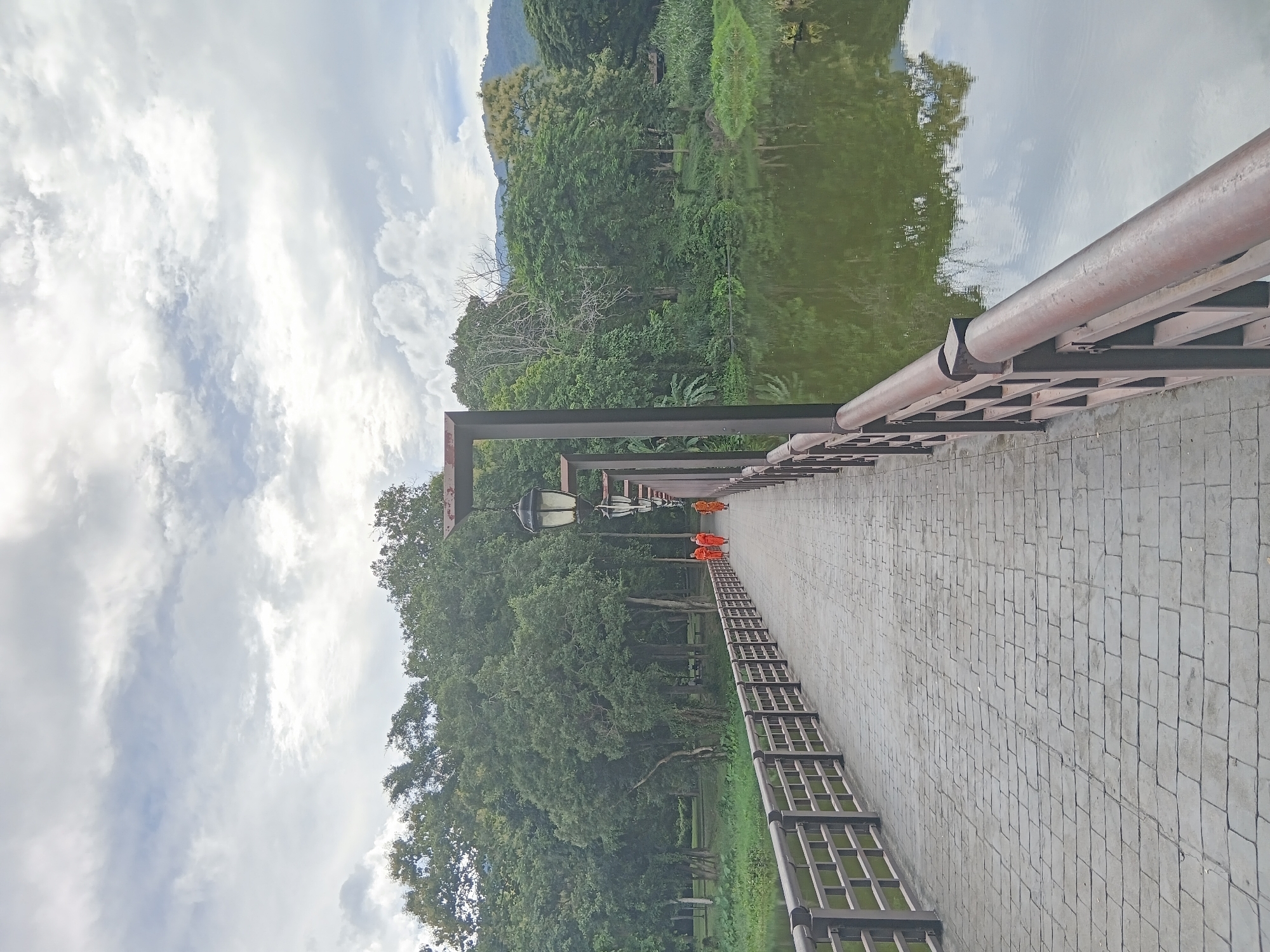 Three Buddhist monks in saffron robes are walking across a paved bridge over a body of water. The bridge has ornate wooden railings and lampposts. Lush green trees line the far bank of the water, with mountains visible in the distance under a cloudy sky. The scene appears to be in Chiang Mai City Municipality, Thailand.