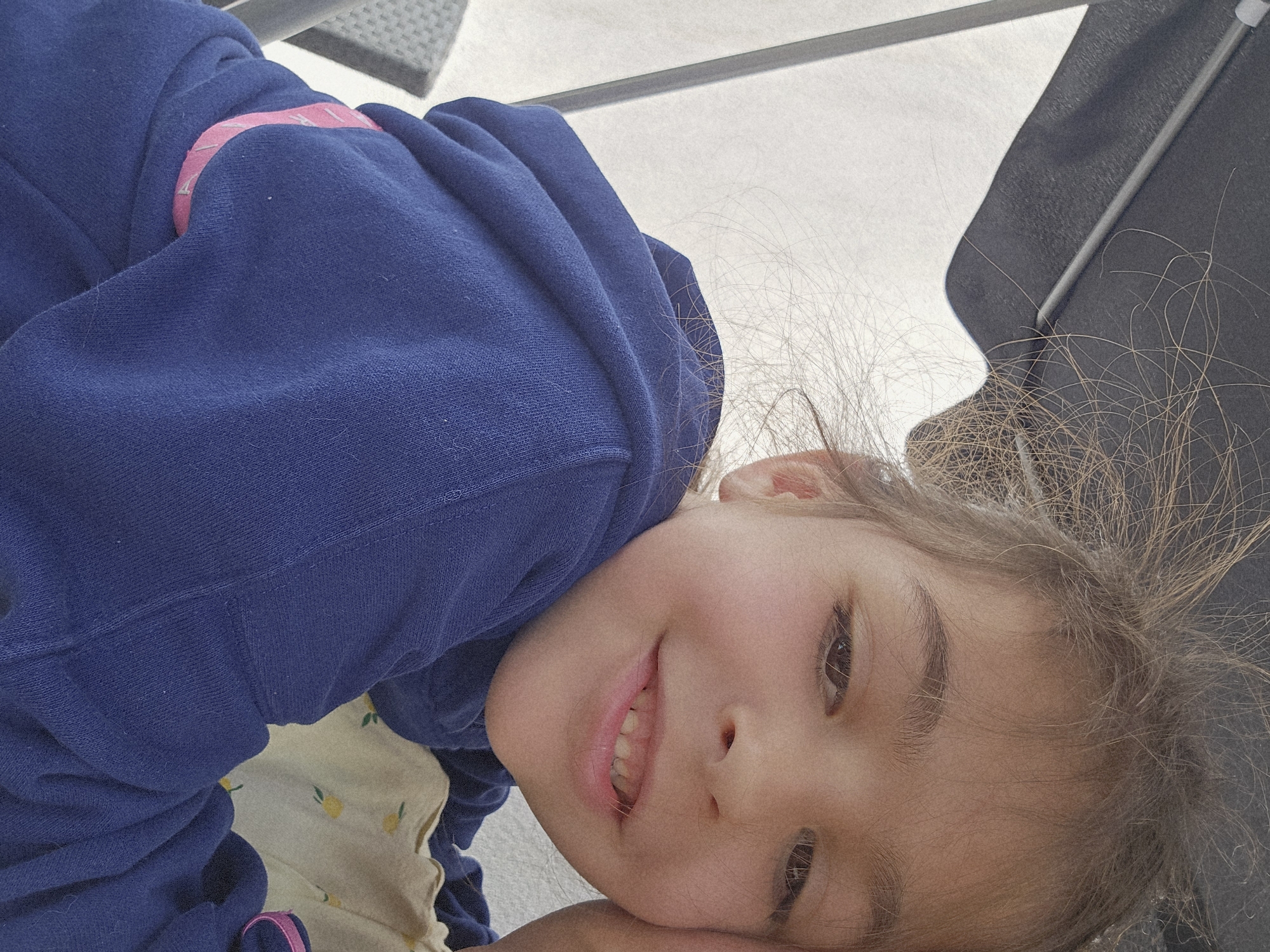 A young girl with brown hair and eyes is lying down. She is wearing a blue hooded sweatshirt with a pink accent and a light-colored shirt with a yellow pattern. The background includes a gray surface and what appears to be a section of an umbrella. The location cannot be determined to be Zirndorf, Germany.