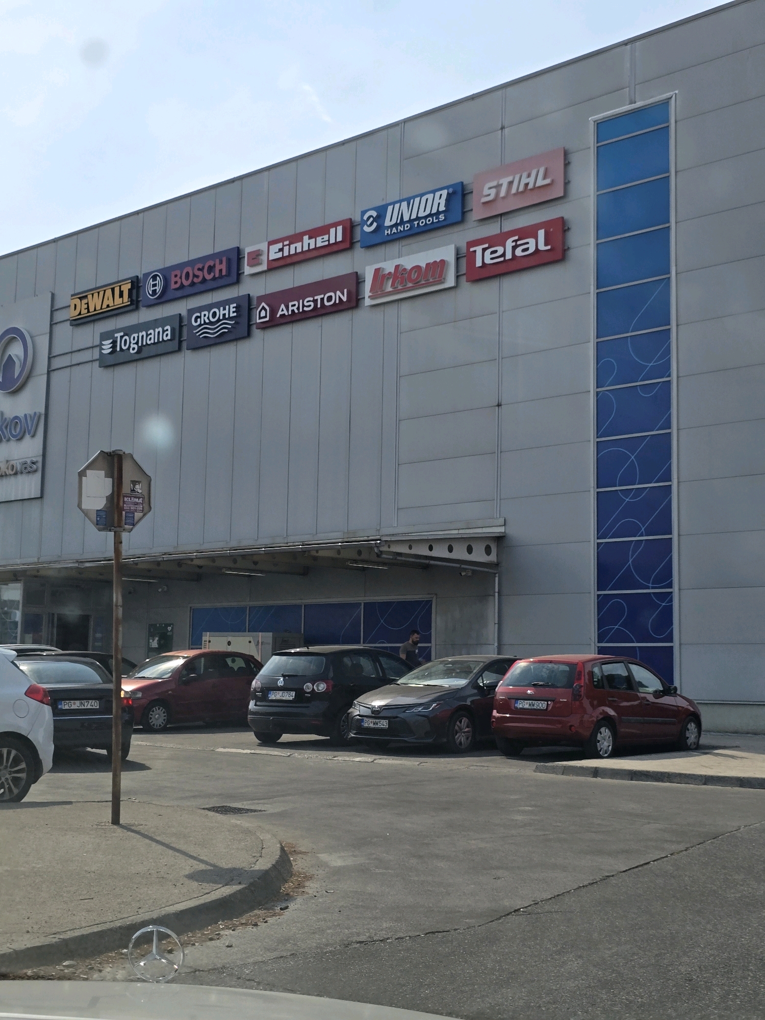 The image captures a storefront facade of a commercial building, likely a retail center, in Podgorica, Montenegro. The building features a large, grey, metallic exterior, adorned with various brand signs including DEWALT, BOSCH, Tognana, GROHE, Einhell, ARISTON, UNIOR HAND TOOLS, STIHL, irkom, and Tefal. A vertical blue-tinted glass panel with white swirling decorative lines is visible on the right side of the building.
In the foreground, several cars are parked in a lot. A red hatchback is parked on the right, a dark grey hatchback is in the center, and a dark grey sedan is on the left. A white vehicle's rear is partially visible on the far left. A street sign is positioned on a pole in front of the building, partially obscuring some of the signs. A Mercedes-Benz emblem is visible on the hood of a car in the very bottom foreground. One person, a male, is partially visible standing near the entrance of the building on the left. The sky is clear with some light clouds.