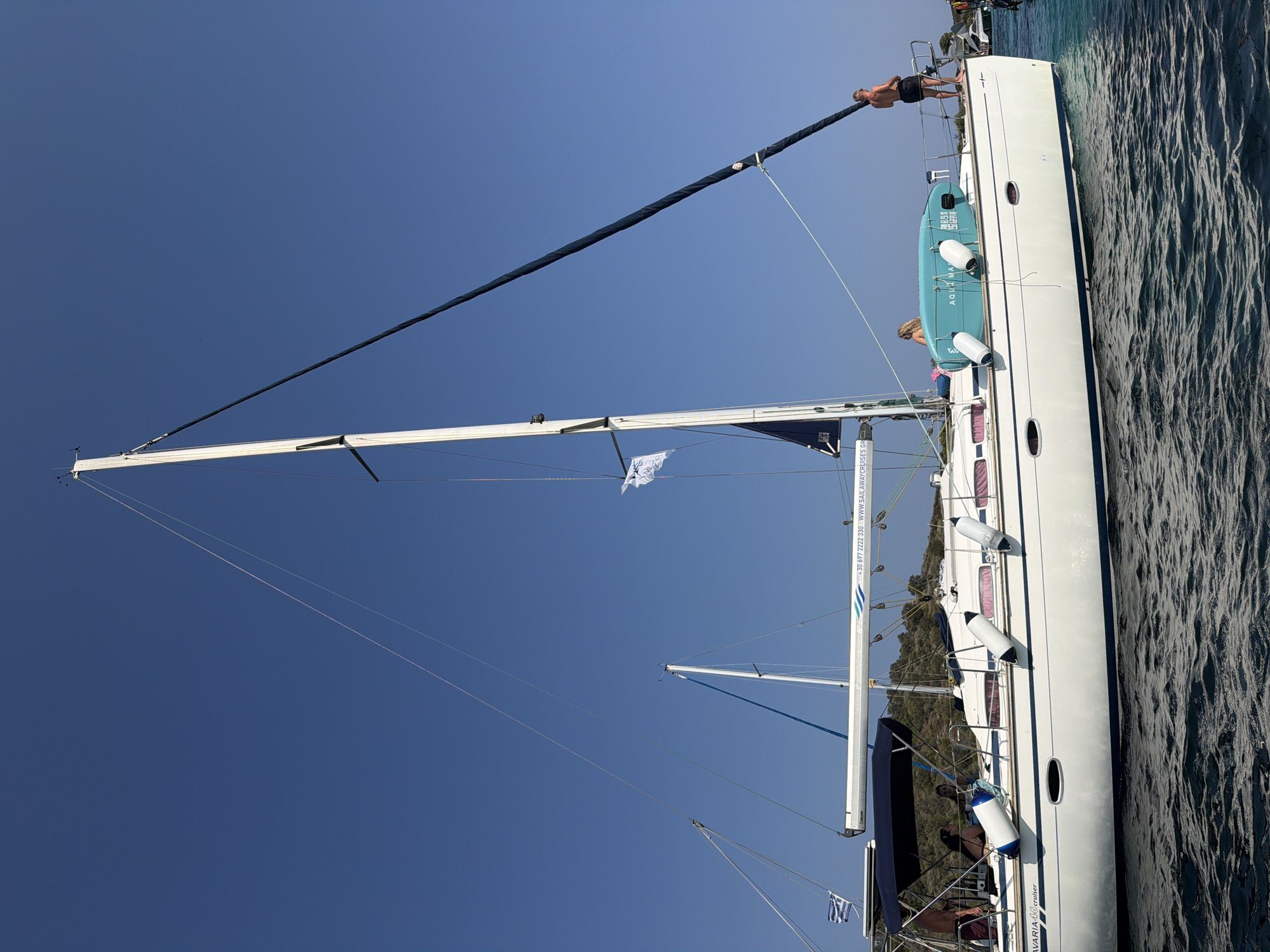A white sailboat with blue trim is anchored in the clear blue water near a small, green, hilly island. The sailboat has a tall white mast and a dark blue furled sail. Several people are on deck. A man in black shorts stands at the stern, looking out to sea. A woman with blonde hair sits near the middle of the boat, facing away from the camera. Another person is partially visible under a blue awning at the front of the boat. Two white fenders hang from the side of the boat. A light blue paddleboard labeled "AQUA MATE" is secured to the deck. The boat's name, "BAVARIA 410 cruiser", is visible on the hull, as well as a phone number and website address: "+30 697 222 3330 WWW.SAILAWAYCRUISES.GR". A Greek flag is attached to the front of the boat. The sky is clear and bright blue.