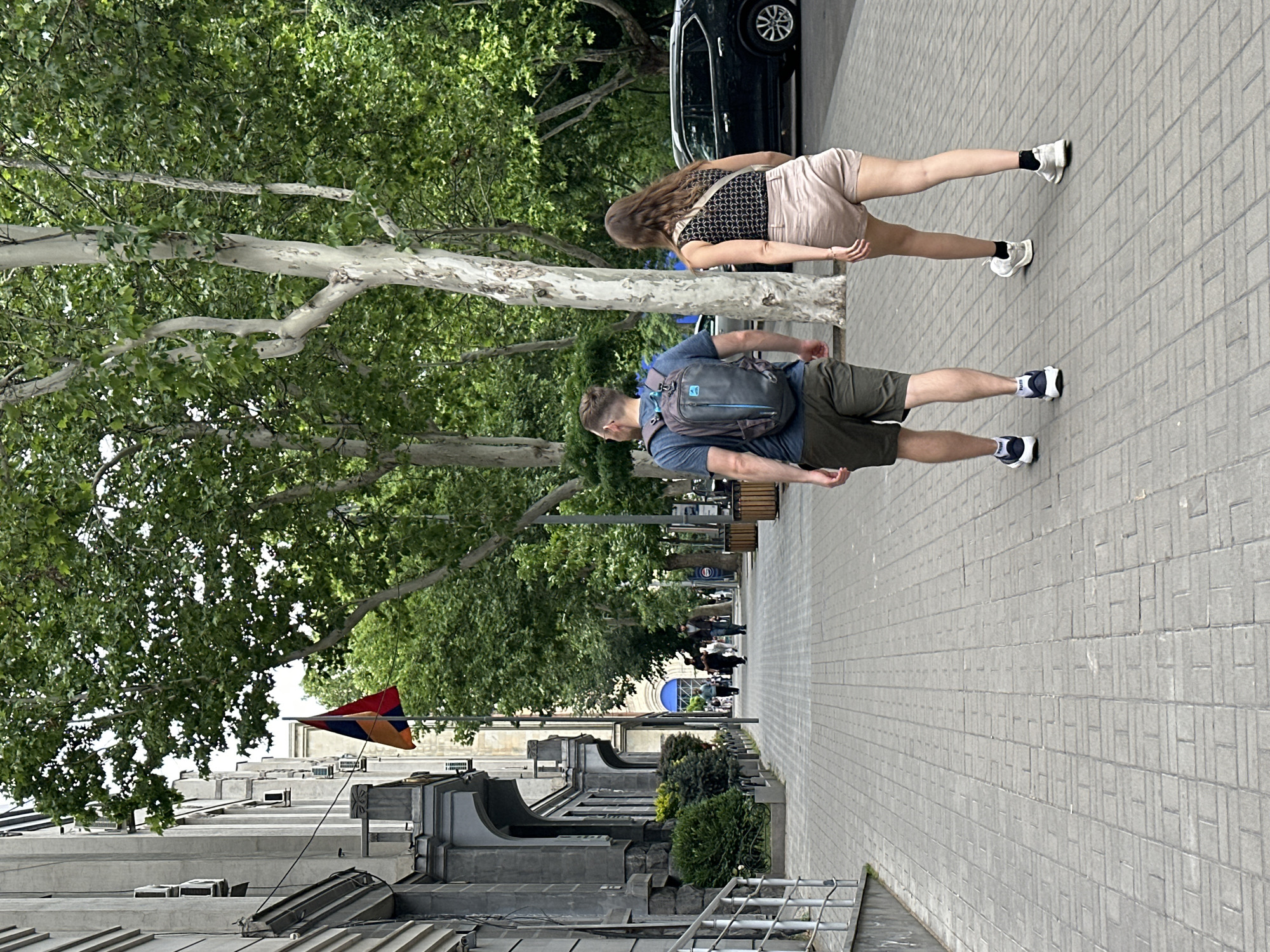 A street scene in Yerevan, Armenia, shows a man and woman walking along a wide paved sidewalk. The man, closer to the camera, wears a blue t-shirt, khaki shorts, a gray backpack with blue accents, and dark and white tennis shoes. The woman, walking slightly ahead, has long brown hair and wears a patterned tank top, tan shorts, and white tennis shoes. They are both walking away from the camera. The sidewalk is lined with trees on one side and buildings on the other. The Armenian flag is visible atop one of the buildings. In the background, other pedestrians can be seen walking along the sidewalk. A dark car is parked on the street next to the sidewalk.
