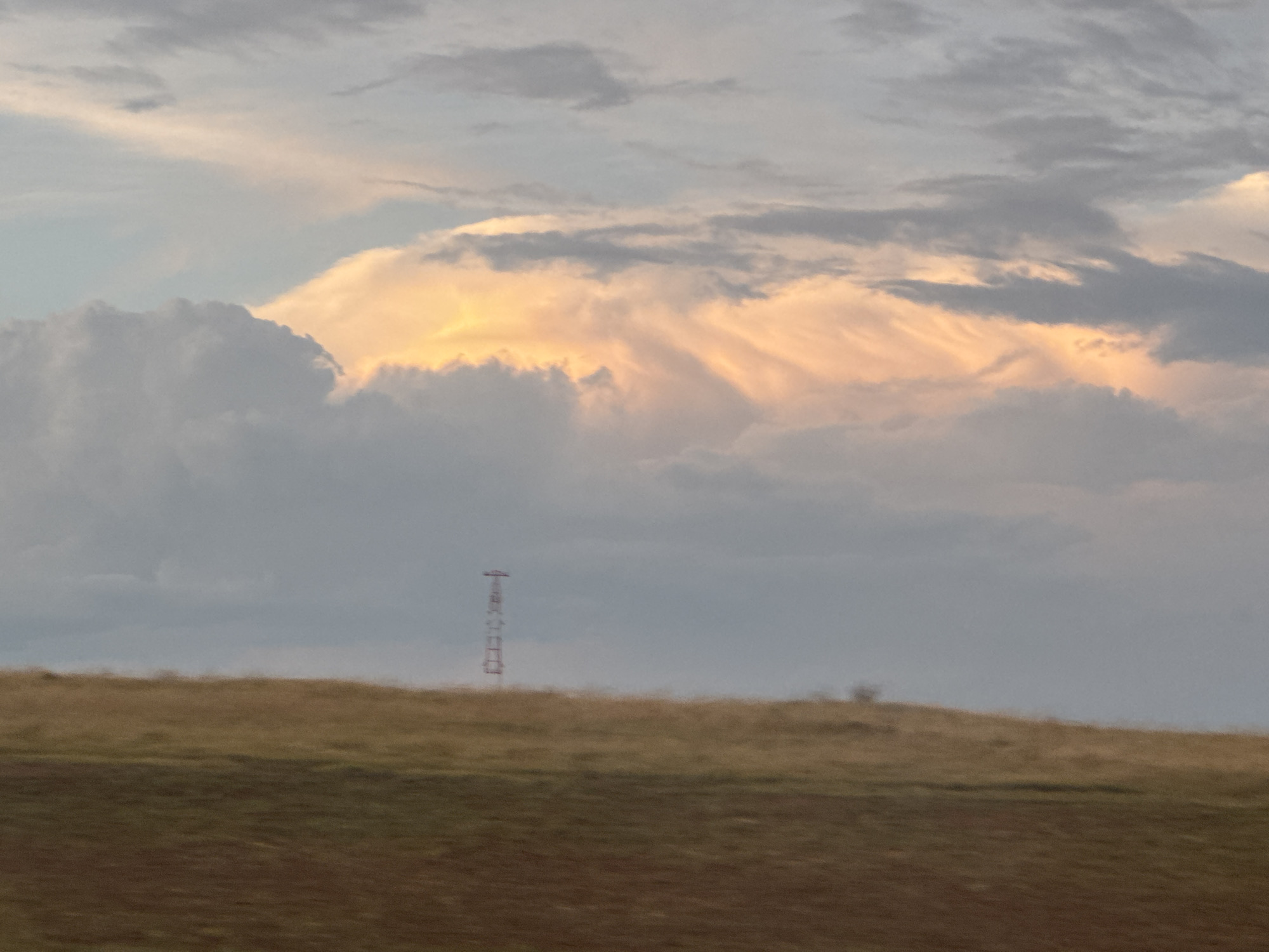 The image depicts a wide, undulating landscape under a dramatic sky. The foreground is divided into two distinct horizontal bands: a darker, reddish-brown area at the very bottom, likely tilled earth, transitions into a lighter, golden-brown expanse of dry grass or low vegetation. In the middle ground, a series of rolling hills are visible, their contours softened by distance and possibly atmospheric conditions.
Dominating the background and upper portion of the image is a turbulent sky filled with various cloud formations. Large, gray, and white cumulus clouds are prominent on the left, while the center and right feature lighter, more ethereal clouds illuminated with warm, peachy-orange hues, suggesting either sunset or sunrise light breaking through. The sky transitions to a pale blue-gray at the top.
A slender, metallic tower, possibly a radio or transmission tower, stands vertically on the horizon, slightly left of the center. It appears to be a red and white lattice structure. A few scattered, indistinct trees or shrubs can be discerned on the higher parts of the hills.
The overall impression is one of natural beauty, with the striking interplay of light and shadow in the clouds contrasting with the muted earth tones of the landscape. The presence of the man-made tower introduces a subtle element of human presence within the otherwise natural setting. The image is not specific to the city of Syrt, Russia.