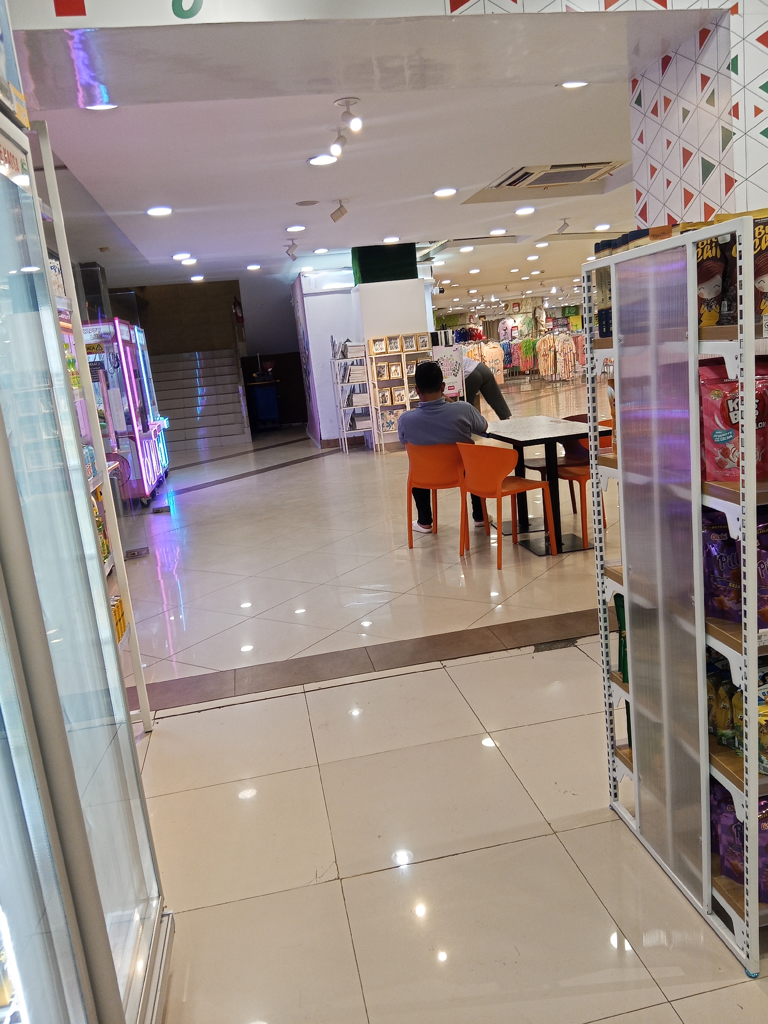 The image depicts an indoor scene, likely a mall or a large retail establishment, in Lubuklinggau, Indonesia. In the foreground, a person is seated at a table with orange chairs. Another person, partially visible, is standing near the table. To the right, shelves stocked with packaged goods are displayed. In the background, there are escalators leading to an upper level, and clothing racks are visible further into the store. The floor is tiled, and the environment is well-lit by overhead lights and some colored lighting from electronic devices in the distance, such as a claw machine. The overall impression is that of a commercial area with shoppers and merchandise.