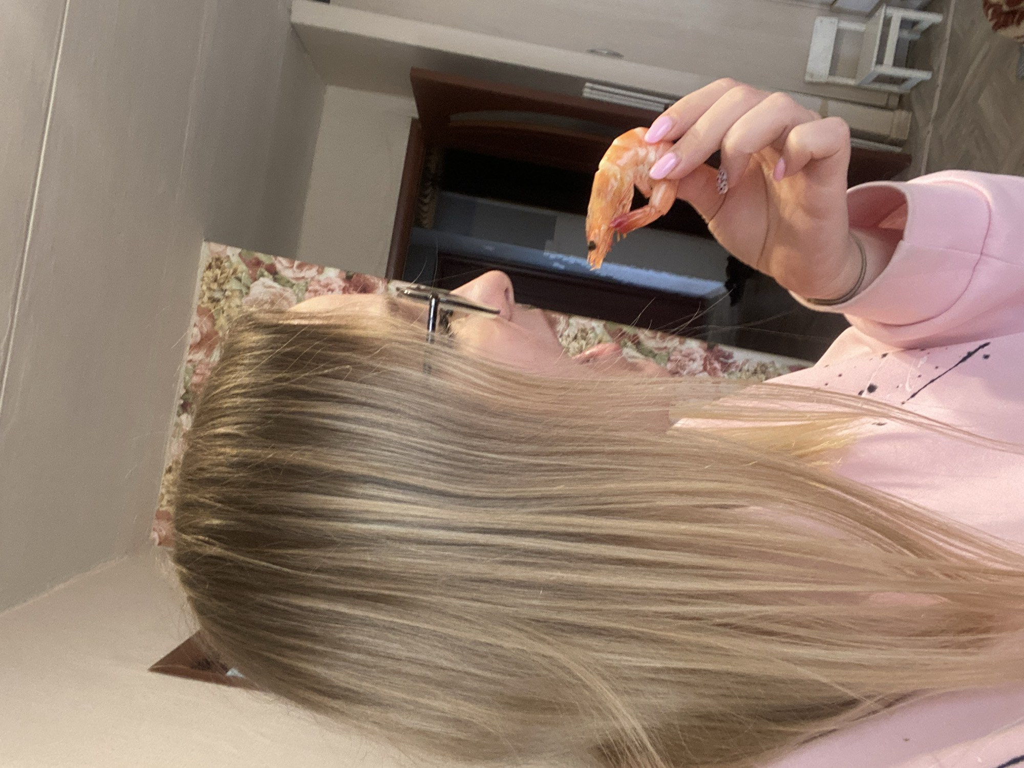 The image shows a person with long, blonde hair lying on a surface with a floral pattern. The person is holding a cooked shrimp near their face. They are wearing glasses and a pink sweatshirt with black paint splatters. The background includes a cabinet or shelf and what appears to be a white structure, possibly a small table or shelf unit. There is no verifiable information to place this image in Kauchuk Stantsiya, Russia.