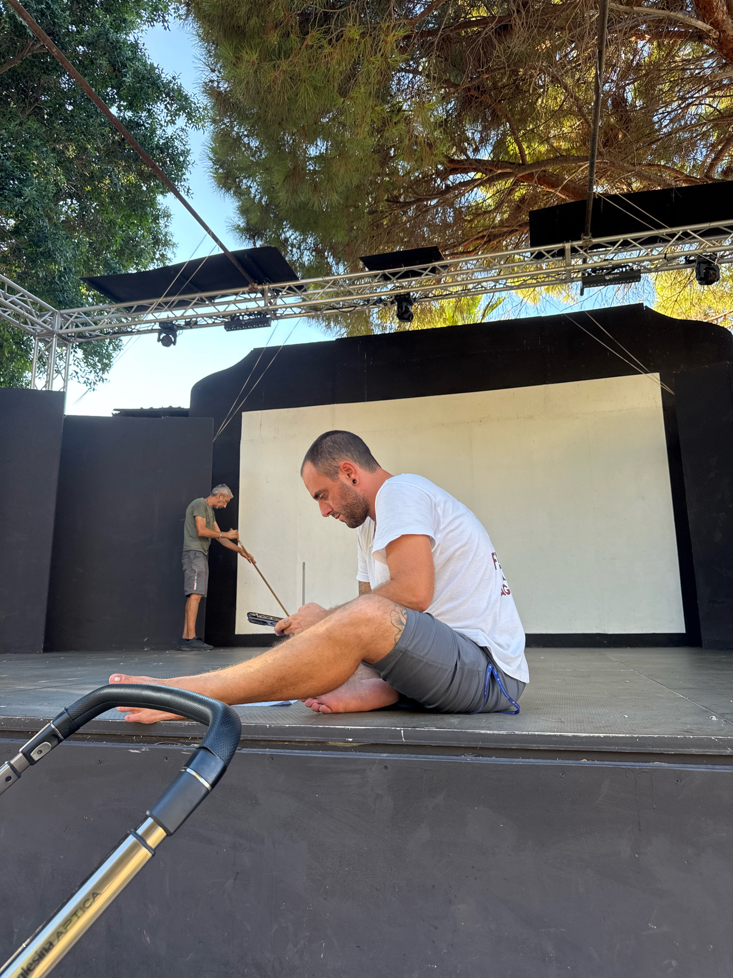 The image depicts an outdoor stage in Isola delle Femmine, Italy, with a white backdrop bordered by black panels. In the foreground, a barefoot man wearing a white t-shirt and grey shorts is sitting on the stage, looking down at a small black object in his hands. To the left, further back on the stage, another man in a green t-shirt and grey shorts is standing and appears to be holding a long stick or pole. The stage is set up outdoors, with trees and sky visible in the background, suggesting a warm, sunny day. A portion of a stroller is visible in the lower left corner of the image.