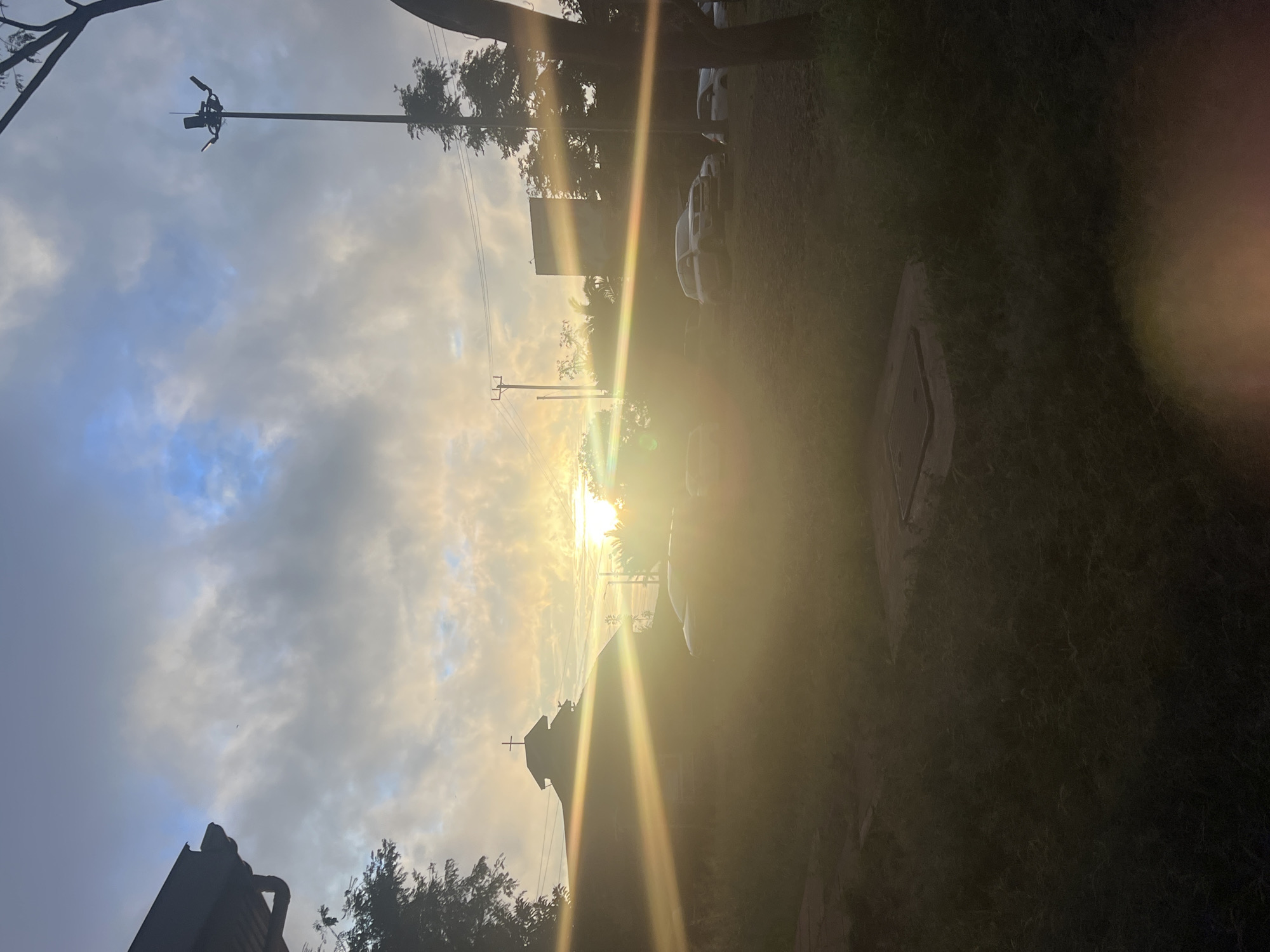 The image shows a daytime scene with the sun shining brightly, creating a lens flare. A church is silhouetted against the sun, suggesting a possible religious setting. Utility poles with wires are visible against a cloudy sky, and cars appear to be parked along a roadside. The environment looks like a suburb in Nairobi, Kenya.