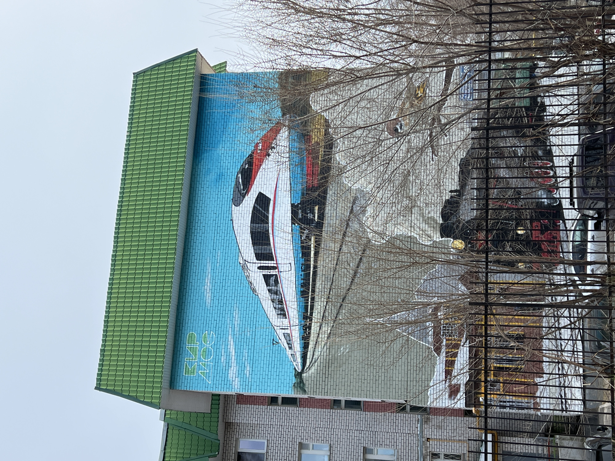 The image depicts a large mural on the side of a building in Kazan, Russia. The mural features a high-speed train painted against a blue sky background, with the text "ЭЛИС МОС" vertically aligned next to it. The building has a green tiled roof and is constructed of white bricks. The mural is partially obscured by tree branches and a metal fence or railing. The sky is overcast. The mural also features a second train drawn into the brickwork of the building.