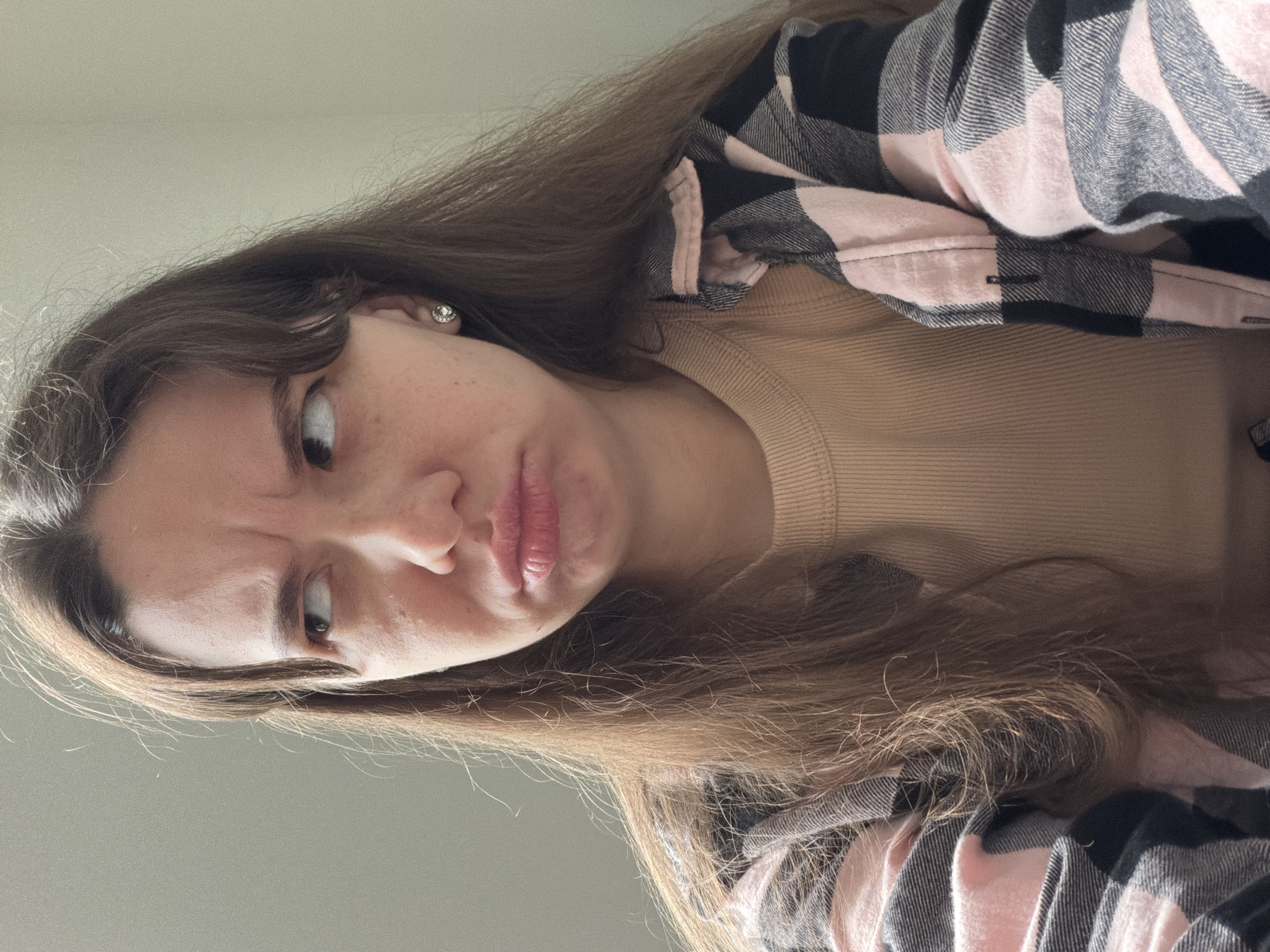 A young woman with long brown hair is looking to the left with a pouty expression. She is wearing a tan ribbed crewneck shirt underneath an open pink, black, and gray plaid shirt. She has a diamond stud earring in her visible ear. The background is a plain, light-colored wall. The setting does not provide any specific details about the location within the city of Kyiv, Ukraine.