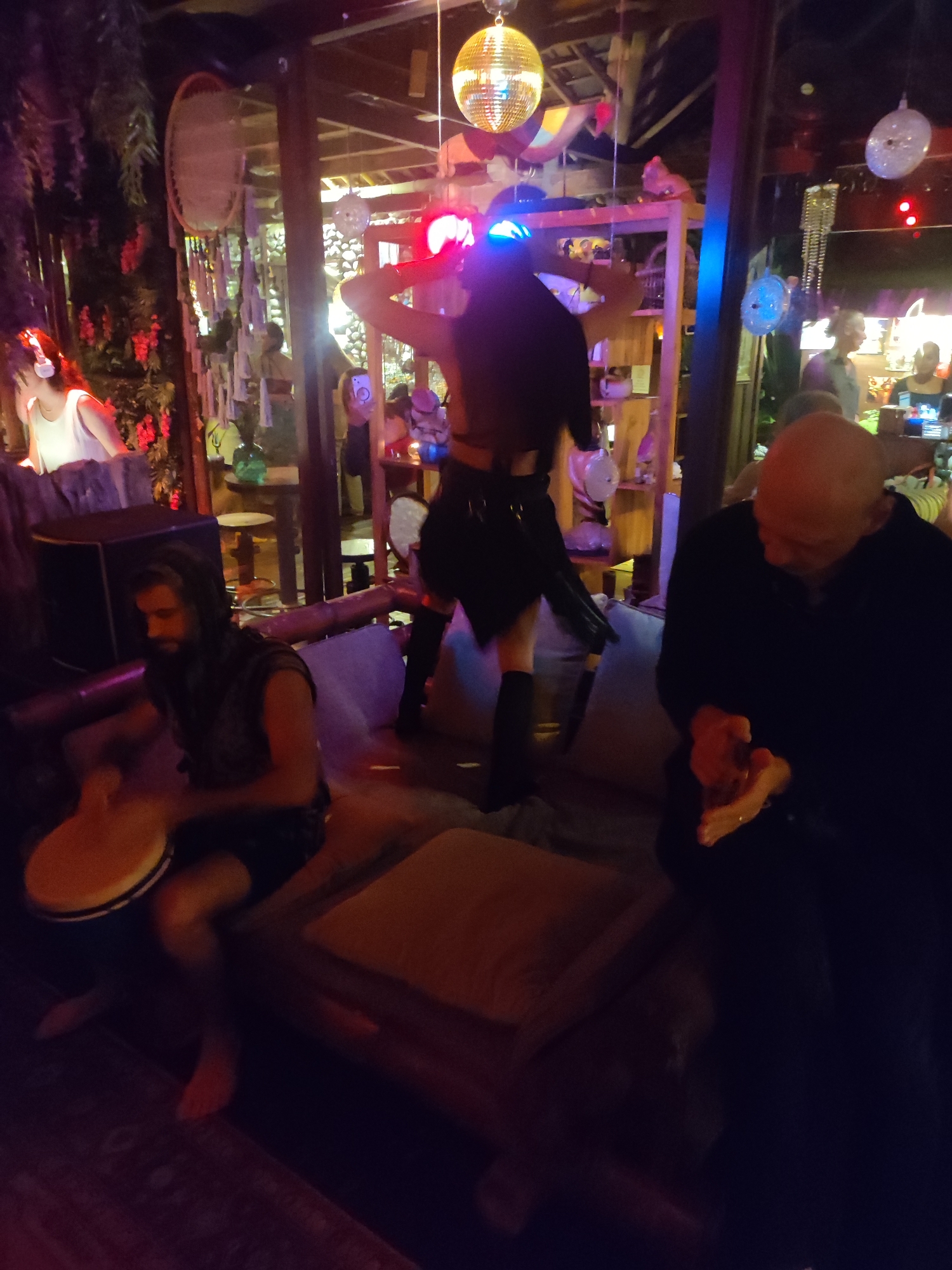 The image depicts an indoor nightlife scene, possibly a bar or lounge, in Lokaserena, Indonesia. Dim lighting is evident. A DJ with illuminated headwear stands on a raised platform in the center of the frame, apparently dancing with hands raised. A man plays a drum, seated on a cushioned surface to the left. To the right, a bald man is seated, looking down and rubbing his hands together. Other patrons can be seen in the background, engaging in conversation. Decorative elements include hanging lights, greenery, and other adornments. The overall atmosphere appears to be relaxed and celebratory.