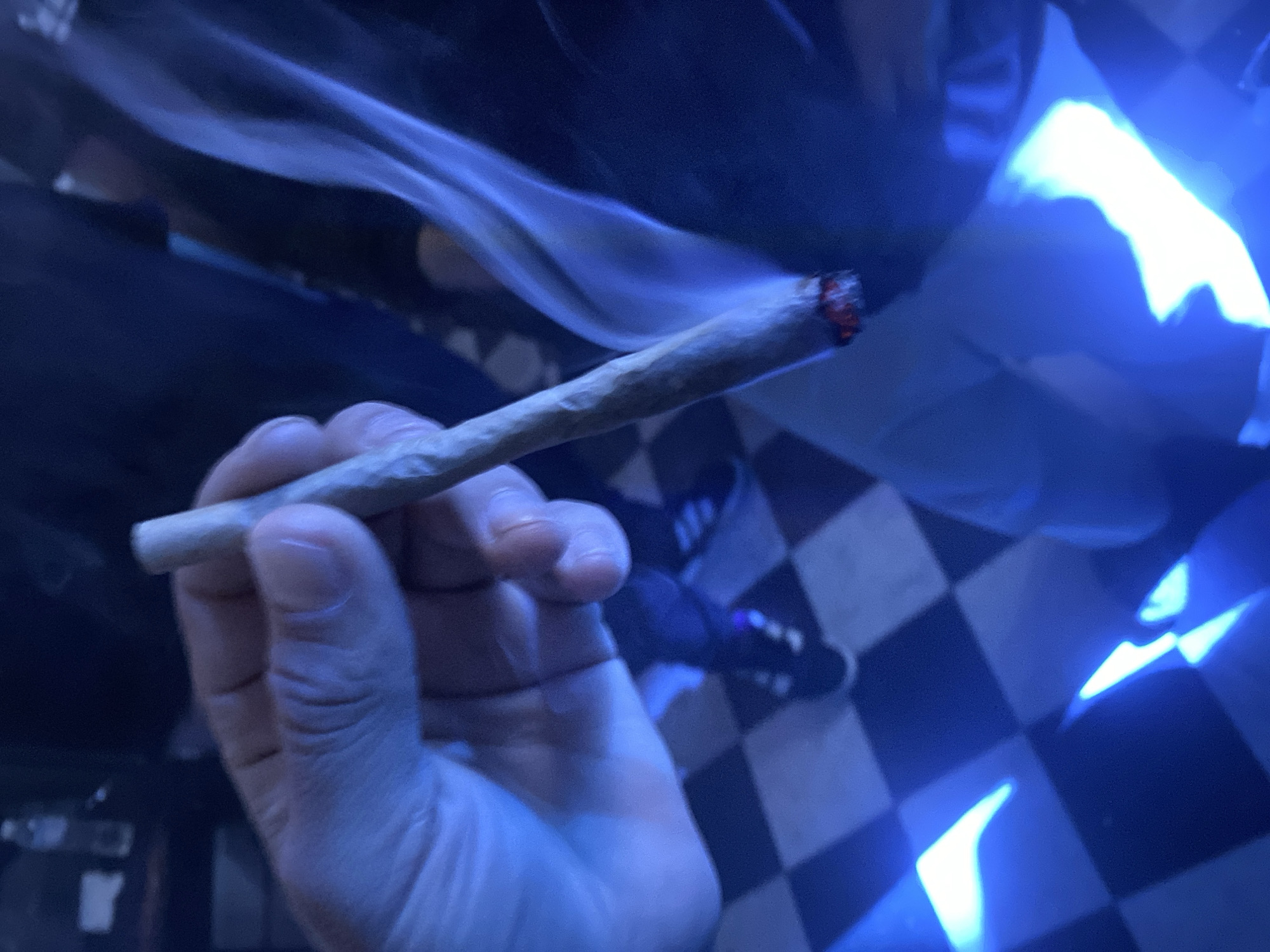A hand holds a lit cigarette or joint indoors. Smoke curls upward from the burning end. The floor is a black and white checkerboard pattern. Parts of shoes and legs are visible in the background. The scene is lit with a blue-toned light. The location and specific details about people are not discernible based on this image.