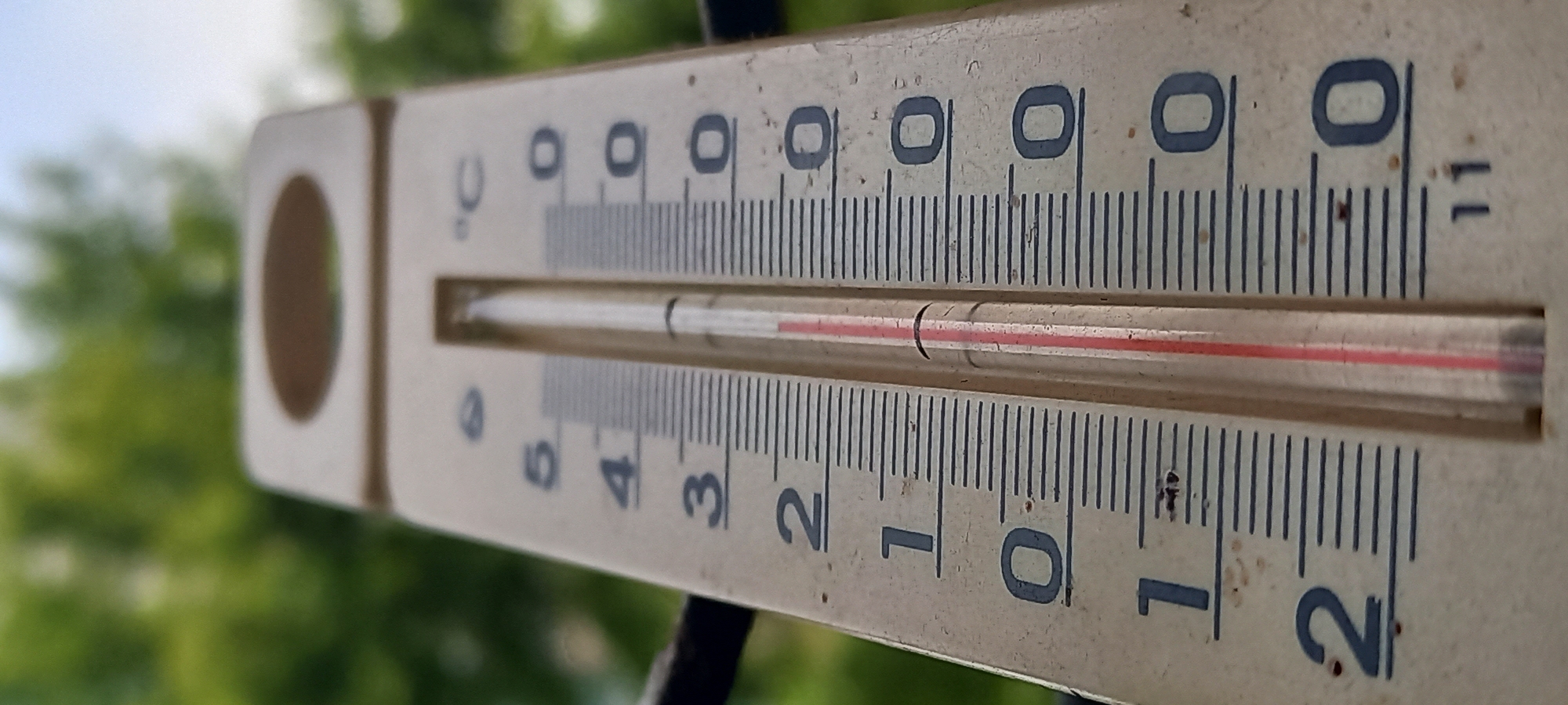 An analog outdoor thermometer, positioned vertically against a blurred background, displays a temperature of +0.7 degrees Celsius. The device features an off-white, weathered plastic or metal casing with a transparent glass tube containing a red liquid column. The Celsius scale is clearly visible, with prominent numerical markings on the left side indicating units from at least -2 to +5. Finer graduations between integer marks represent 0.1-degree increments. The red liquid's meniscus aligns precisely with the seventh minor mark above the '0' (zero degrees Celsius) line. The casing exhibits minor surface dirt and wear, indicative of outdoor exposure. The background consists of heavily blurred green foliage on the left and a lighter, indistinct sky-like element on the right, confirming an exterior setting. No individuals or specific actions are depicted. The scene is located in Kryvyi Rih, Ukraine.