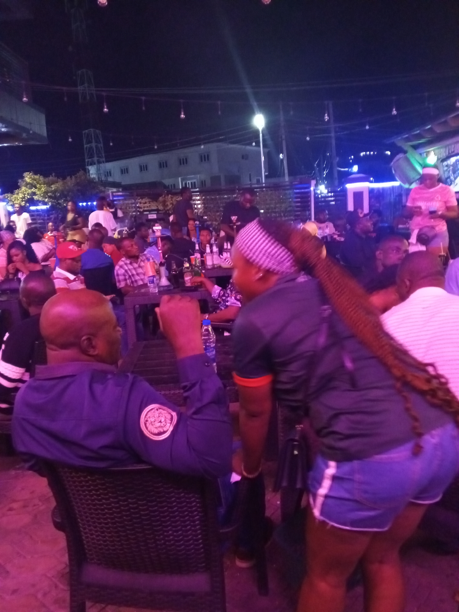 The image depicts a vibrant night scene at an outdoor bar or lounge in Lokoja, Nigeria, filled with people socializing under artificial lighting. The dominant hues are purple and pink from various light sources, creating a lively atmosphere.
In the immediate foreground, a man with a bald head is seated with his back to the viewer, wearing a dark blue long-sleeved shirt that features a circular emblem or patch on his right sleeve. Beside him, a woman stands, seen from behind, notable for her long, dark braided hair extending down her back. She wears a striped headband, a dark short-sleeved top with an orange band on the sleeve, and light blue denim shorts with white stripes down the sides.
The midground is densely populated with individuals gathered around tables, some seated and others standing. Many are engaged in conversation, and various bottles, glasses, and what appear to be shisha pipes are visible on the tabletops. The crowd consists of a mix of men and women dressed in casual attire.
In the background, more patrons can be seen, along with elements of the surrounding environment. A tall, skeletal metal tower structure and multi-story buildings with illuminated windows stand against the dark night sky. Overhead, strings of glowing lights contribute to the festive ambiance, complemented by a bright street lamp in the distance. The overall impression is that of a bustling recreational spot enjoying an evening social gathering.